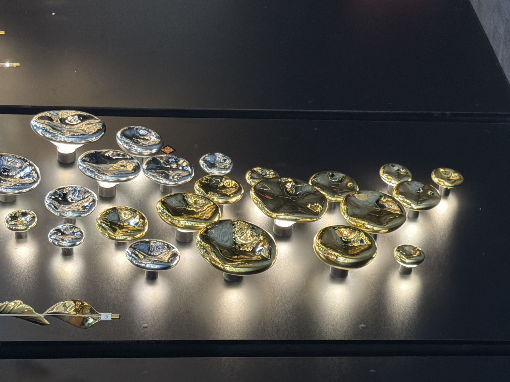 This image shows a collection of abstract, reflective wall sconces that appear to be made of polished metal with gold and silver tones. The sconces are arranged in a vertical cluster on a dark, textured wall. Each sconce has an irregular, organic shape, resembling pebbles or drops of liquid. They are illuminated from behind by a soft, warm light, creating a dramatic effect that highlights their reflective surfaces and the textures of the wall. Some sconces are larger and more prominent, while others are smaller and set further back. The overall impression is one of modern, artistic interior design, possibly in a commercial or exhibition space. There are no people visible in the image. The provided information does not specify a location in Moscow, Russia.