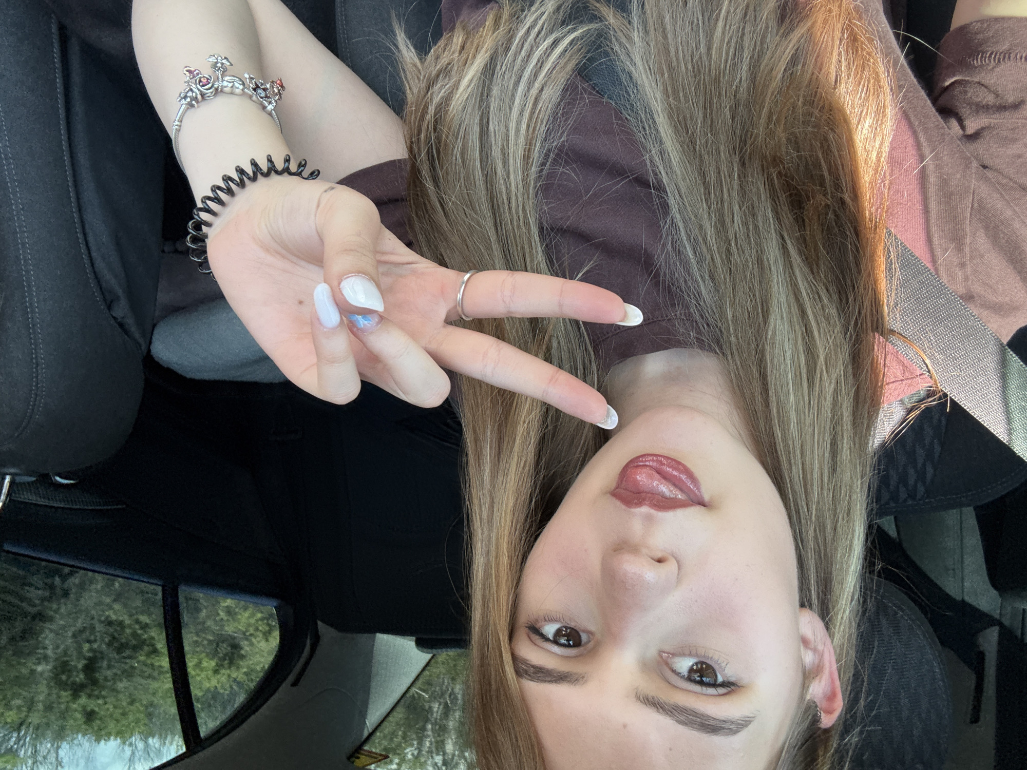 A young woman is in a car, likely a passenger, as indicated by the seatbelt across her shoulder. She's holding up her hand in a "peace" or "victory" sign. Her nails are painted white with some decoration on one nail. She's wearing rings and bracelets on her hand/wrist. The interior of the car is visible, including the seat and window. Outside the window, trees are visible. The photo does not provide enough context to determine the exact location in Levski, Bulgaria.