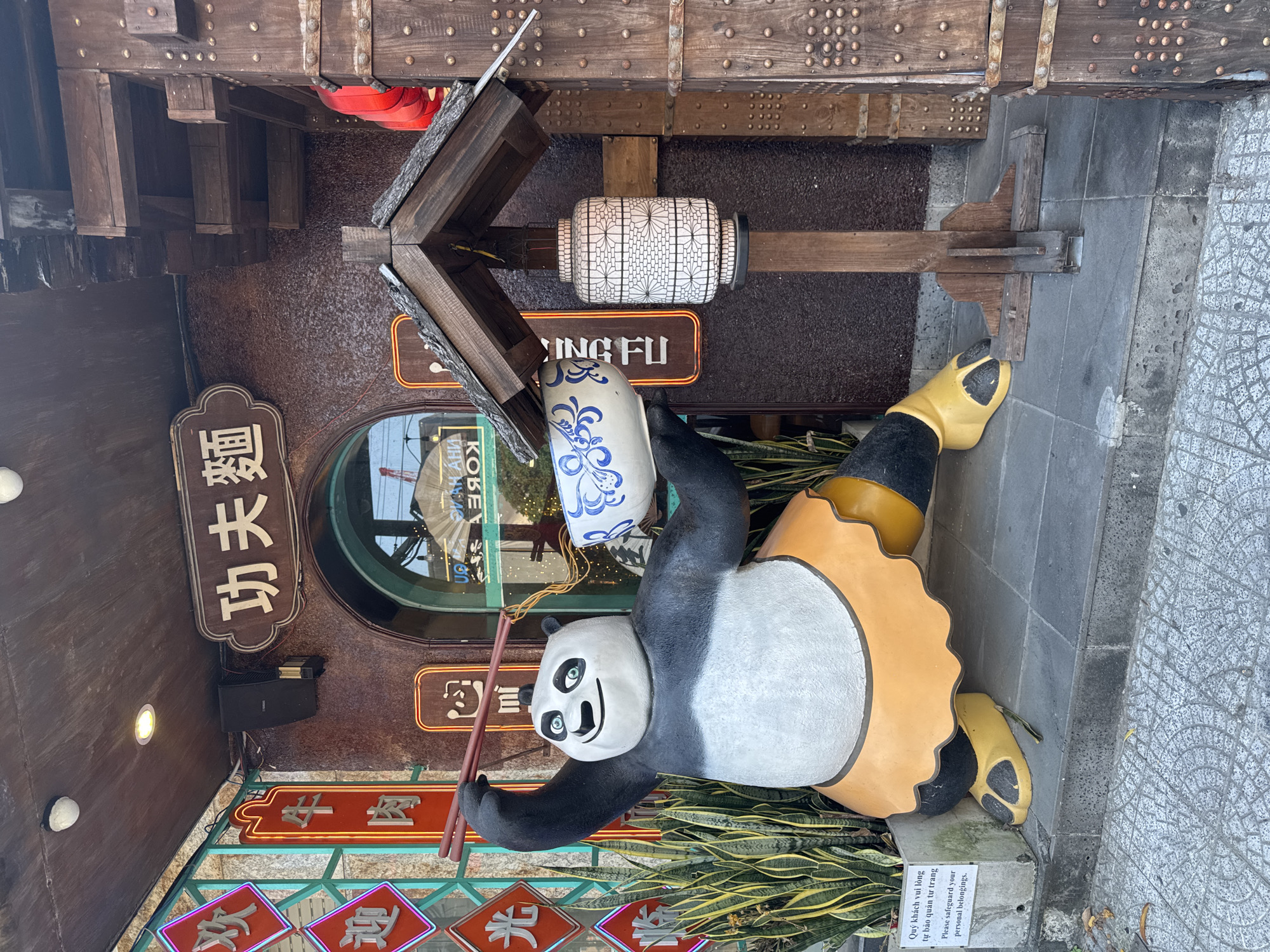 The image shows a statue of a panda holding a ceramic bowl and chopsticks, positioned in front of a restaurant in Đà Nẵng, Vietnam. The panda statue is laying on its back near some snake plants. The restaurant's storefront features a mix of Chinese characters and English text. The signs above the restaurant entrance say "牛肉", "欢迎光", "功夫麵", and "SING FU". A sign below the panda asks patrons to "Please safeguard your personal belongings". The overall aesthetic incorporates traditional Chinese design elements.