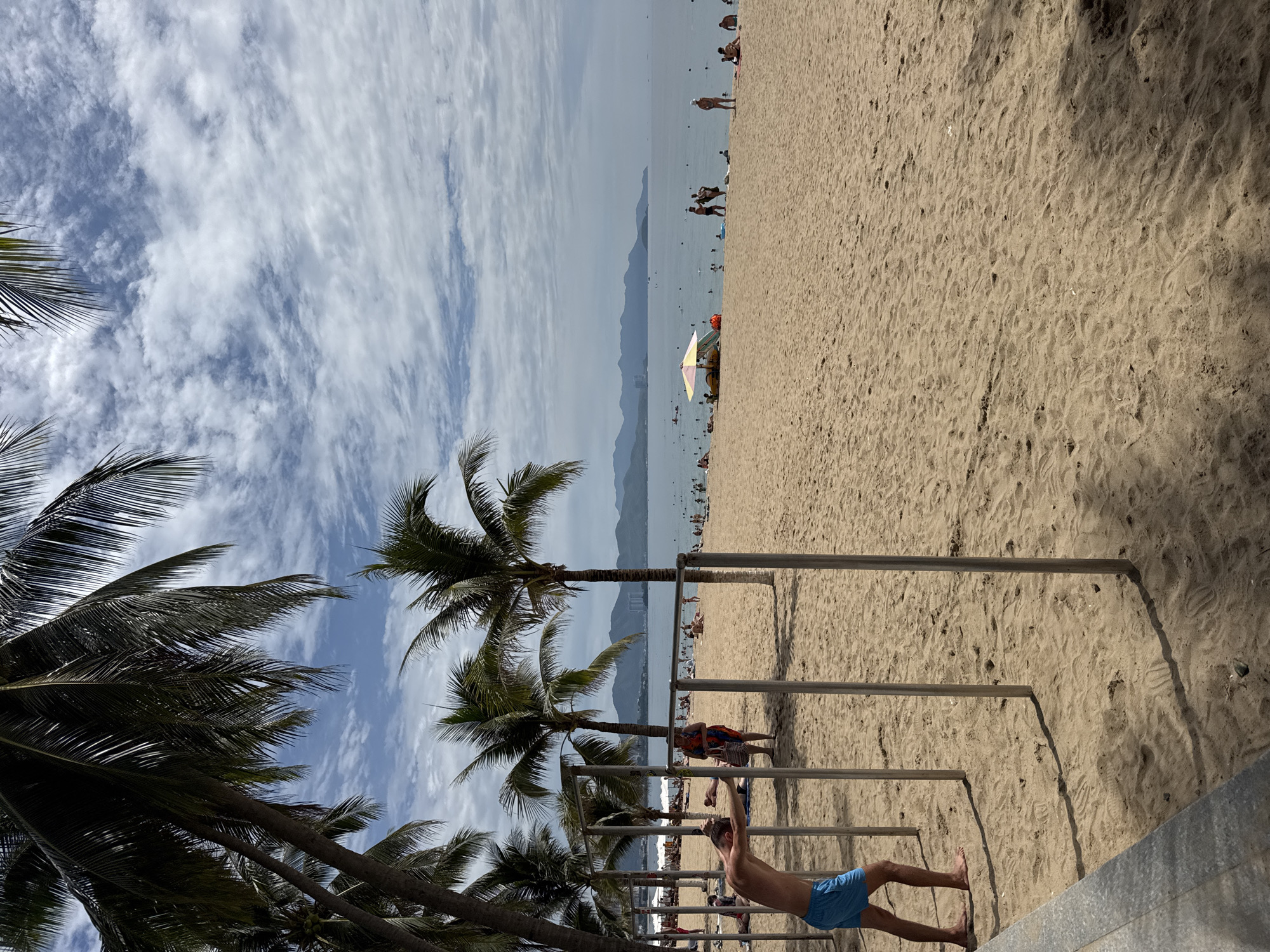 The image depicts a sandy beach with scattered palm trees and a body of water in the background, likely Nha Trang Bay in Nha Trang, Vietnam. The sky is partly cloudy. In the foreground, a man wearing blue shorts is leaning on a horizontal bar, appearing to be exercising. Several metal poles, possibly part of an outdoor gym, are visible. Further back, numerous beachgoers are scattered across the sand and in the water. Some are lying down, others are walking, and some are wading or swimming. Beach umbrellas are visible, providing shade. The coastline stretches into the distance, with mountains visible on the horizon.