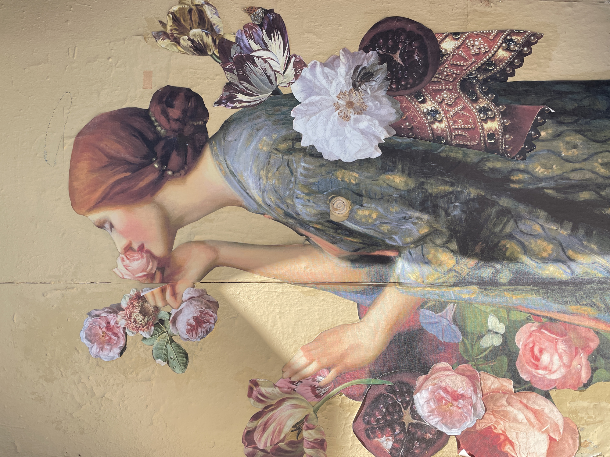 This is a collage artwork depicting a woman with auburn hair pulled back in a bun, smelling a rose. She is wearing a blue and gold patterned robe. Surrounding her are various floral elements, including pink roses, tulips, and a white flower. Pomegranates and a patterned fabric trim are also incorporated into the collage. The artwork is affixed to a textured, pale yellow wall. There is a vertical seam running down the center of the wall, suggesting it is made of two panels. A shadow falls across the woman's body from the upper left. The artwork does not appear to be in Saint Petersburg, Russia, but rather in a studio or gallery setting.