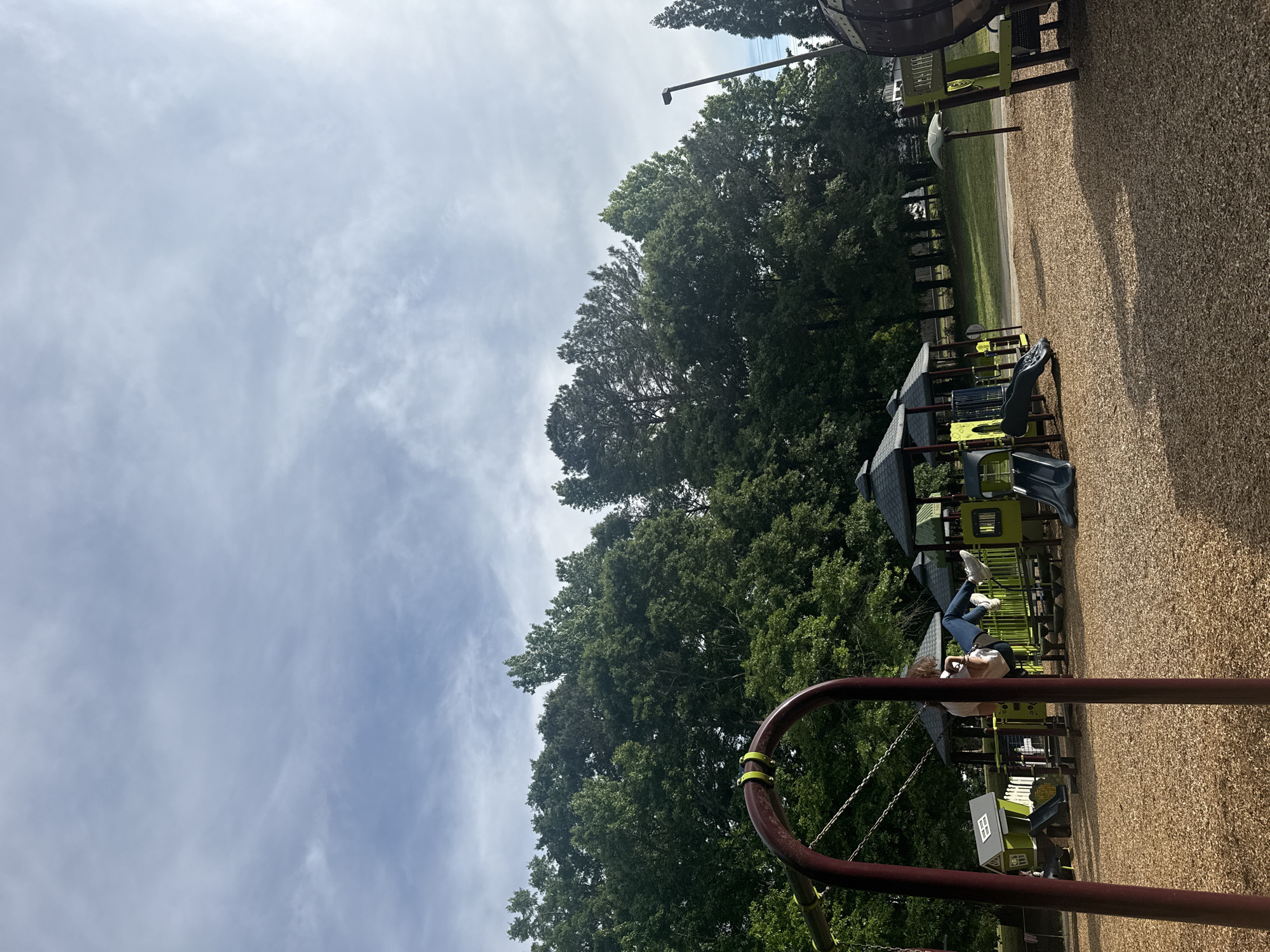 The image depicts a playground scene in Mountain Park, United States of America. A variety of playground equipment can be seen, including slides, structures with roofs, and a climbing frame in the foreground. The ground is covered with a bark-like material. A person is partially visible near the playground equipment, wearing jeans and white shoes. Lush green trees and a cloudy sky form the backdrop of the scene.