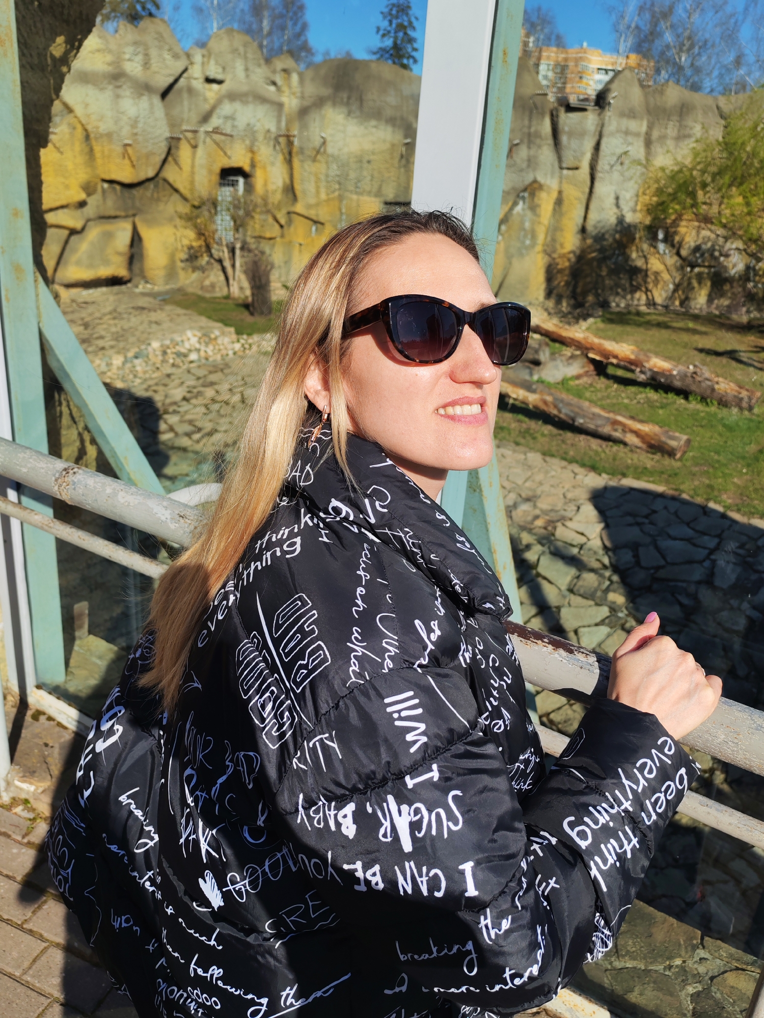 A woman stands on a bridge at the Izhevsk Zoo in Russia. She wears sunglasses and a black puffy jacket with white text. The bridge has a glass panel and metal railings. Behind her are artificial rock structures, some greenery, and a building visible in the distance.