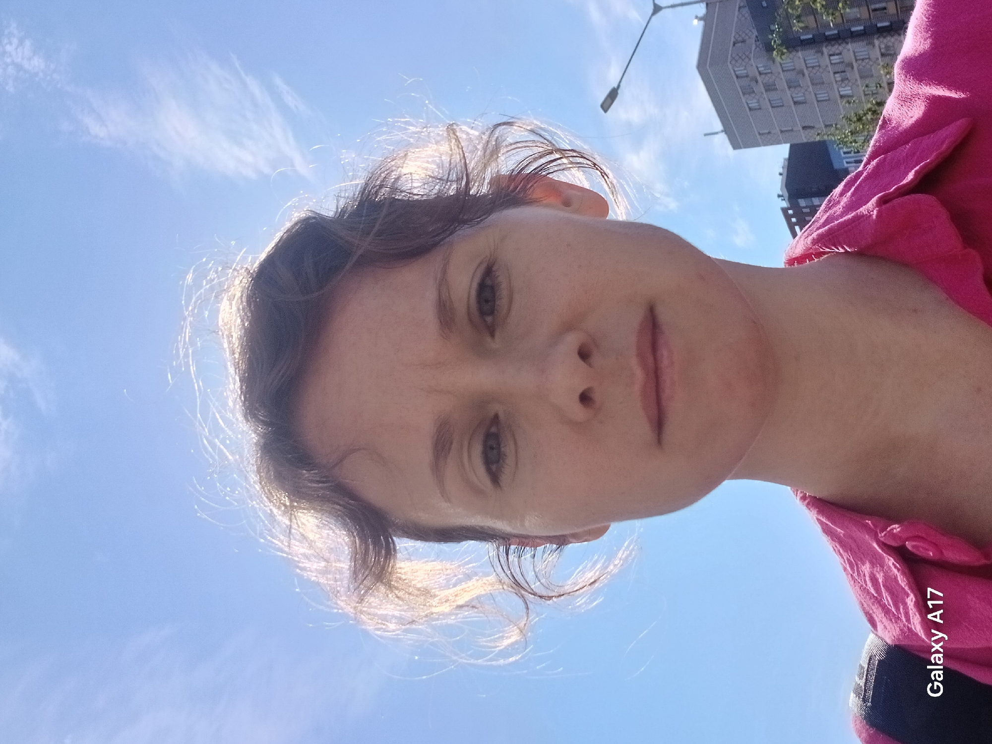The image is a selfie taken from a low angle, showing a woman's face and upper torso against a bright blue sky with wispy clouds. She has light skin, brown hair, and is wearing a pink garment. In the background, there are buildings and a street light. There is a "Galaxy A17" logo visible in the bottom right corner. The location of Yekaterinburg, Russia is unable to be verified from the contents of the provided image.