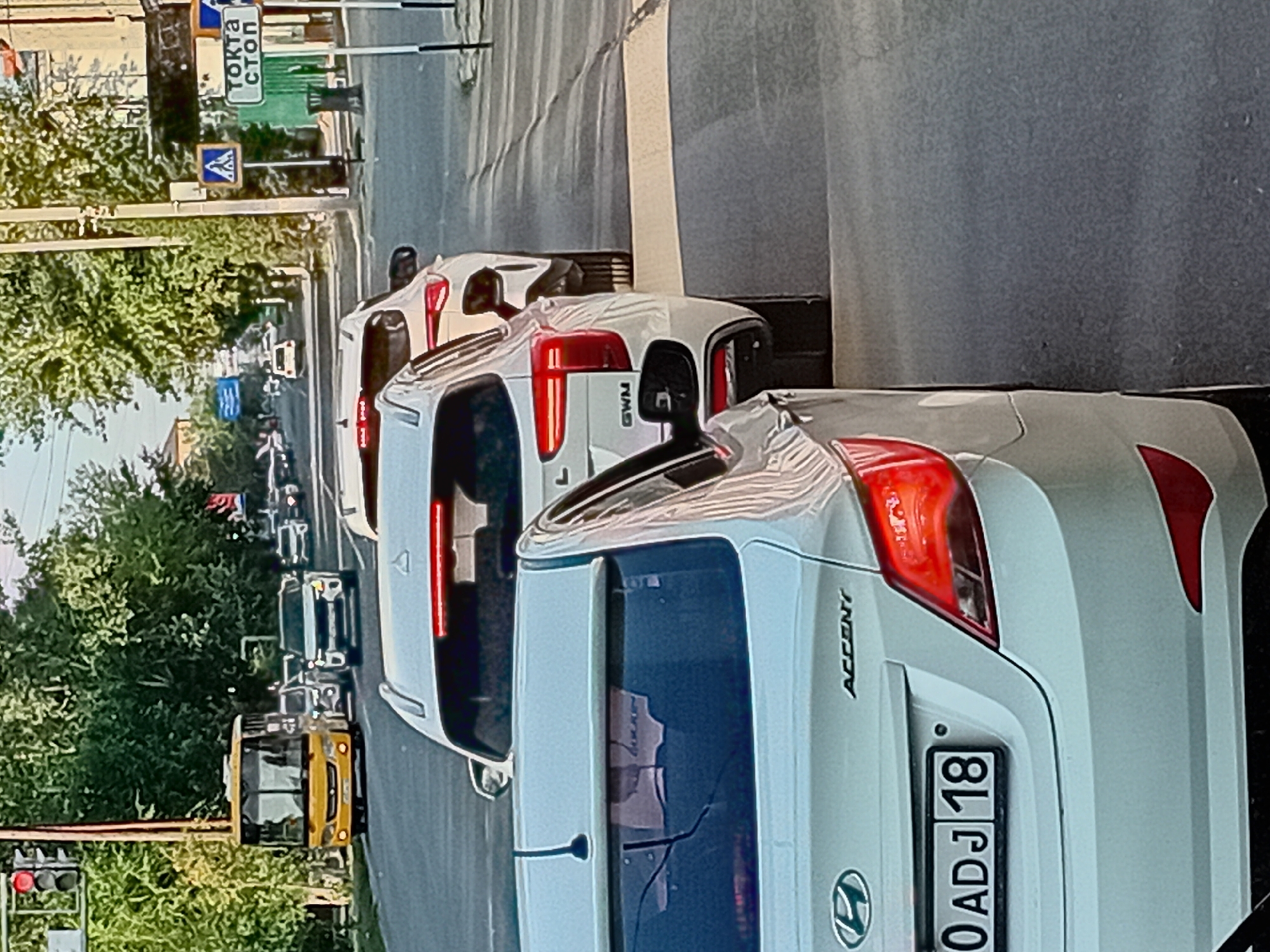 The image shows a street scene in Semey, Kazakhstan, with several cars in traffic. In the foreground, a white Hyundai Accent is partially visible from the rear, displaying the license plate "0 ADJ 18". Further ahead, a white SUV with the model designation "GWM" is in the lane. Behind it, another white SUV and a white sedan are also in traffic. On the left side of the road, a yellow bus is stopped at a traffic light. A pedestrian crossing is visible in the middle ground, marked by yellow and white stripes. Traffic signs are present, including one with a pedestrian symbol and another that appears to be a stop sign. Trees line the street, and buildings can be seen in the background. The overall lighting suggests it is daytime.