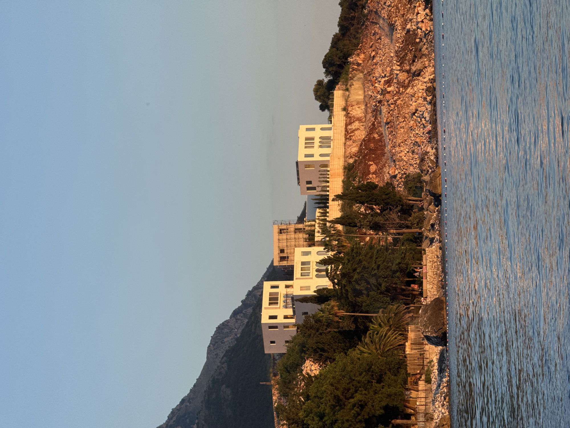 The image shows a waterfront view in Kamenovo, Montenegro. The scene includes the Adriatic Sea on the right, displaying sunlit water ripples. The coastline features a rocky cliff face with vegetation, including trees and bushes, leading up to modern, white buildings built into the hillside. Some of the buildings appear to be under construction, with scaffolding visible. The left side of the image consists of a mountain slope. The sky is clear.