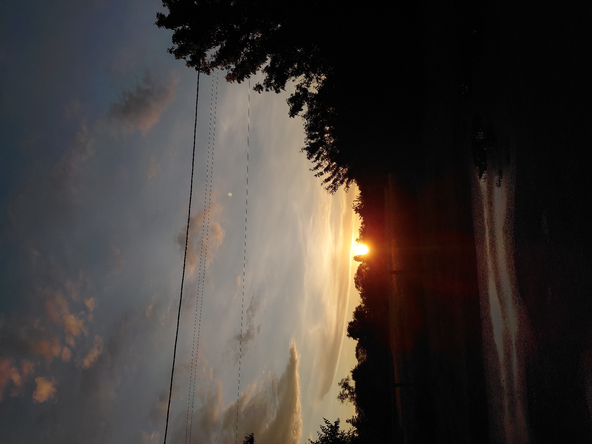 A sunset is captured in a low-angle shot, with the sun low on the horizon and partially obscured by trees. The sky is a blend of dark gray clouds and lighter hues of orange, yellow, and blue. Three power lines run horizontally across the sky. In the foreground, a dark, silhouetted landscape is visible, with trees and vegetation along the horizon. A wet surface, possibly a road or path, reflects the sunset light in the lower portion of the image. A small, pale circle, possibly the moon, is visible in the upper sky. The location is the city of Unknown, Unknown.