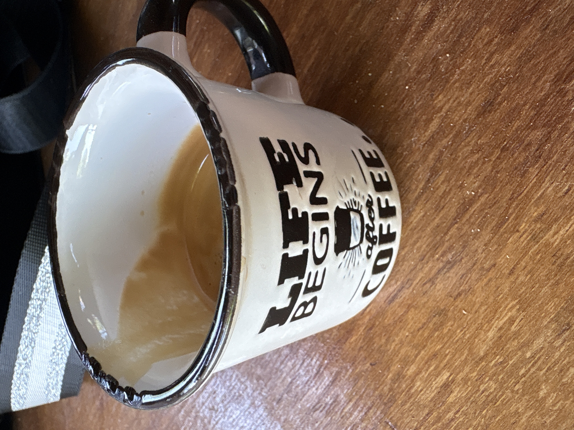 A close-up, overhead shot shows a white ceramic mug with a dark rim and handle, filled with coffee. The mug is decorated with black text that reads "LIFE BEGINS after COFFEE". It sits on a wooden surface, possibly a table. In the upper left corner, a black and white striped fabric is partially visible, along with a black strap. The image is a still life and does not contain any people or specific location information.
