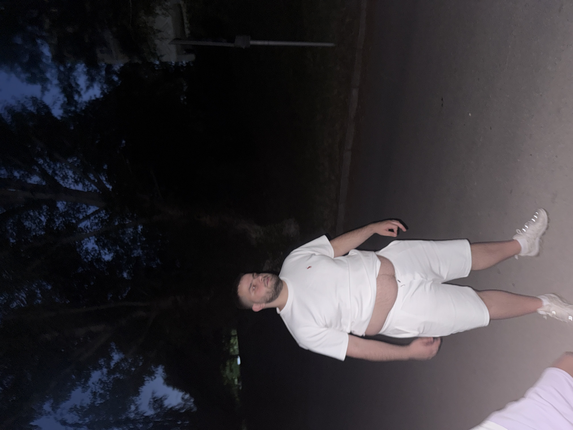 The scene is an outdoor, low-light environment, likely dusk or night, in the city of Blagoevgrad, Bulgaria. A male individual of heavy build with short dark hair and a beard stands on a dark paved surface. He wears a white short-sleeved t-shirt featuring a small red polo horse logo on the left chest, white shorts, white socks, and light-colored athletic shoes with reflective details. His midsection is exposed between the bottom hem of his t-shirt and the waistband of his shorts. He is looking towards the right side of the frame. In the lower-left foreground, a portion of another individual is visible, including a light purple garment and the front of a white shoe. The background consists of dark tree silhouettes against a deep blue-grey sky, indicating ambient light conditions. A thin, dark pole is visible vertically on the far right side of the frame, emerging from a darker, possibly vegetated, area. The ground surface appears to be asphalt or similar paving, with a discernible curb line along the left edge.