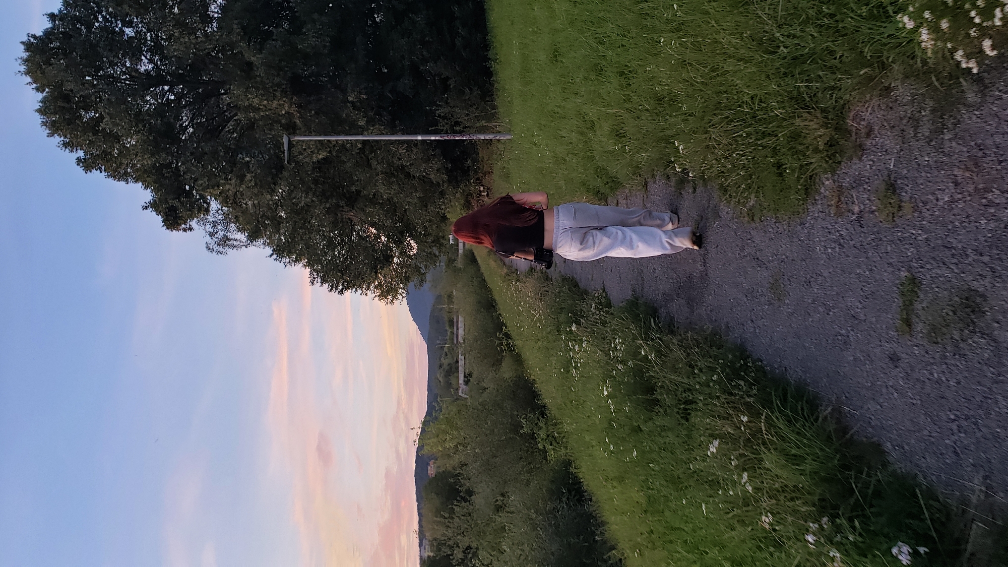 A young woman with long, red hair walks away from the camera on a gravel path. She is wearing a black crop top and light-colored, loose-fitting pants. She is carrying a black camera bag over her shoulder. The path is bordered by lush green grass with small white wildflowers. In the background, there are dense trees and rolling hills under a soft, pink and blue sunset sky. The scene appears to be in a natural or park-like setting, potentially on the outskirts of Prešov, Slovakia, given the hint. A lone street light stands tall on the right side of the path, near a large tree. The overall impression is one of a peaceful evening stroll.