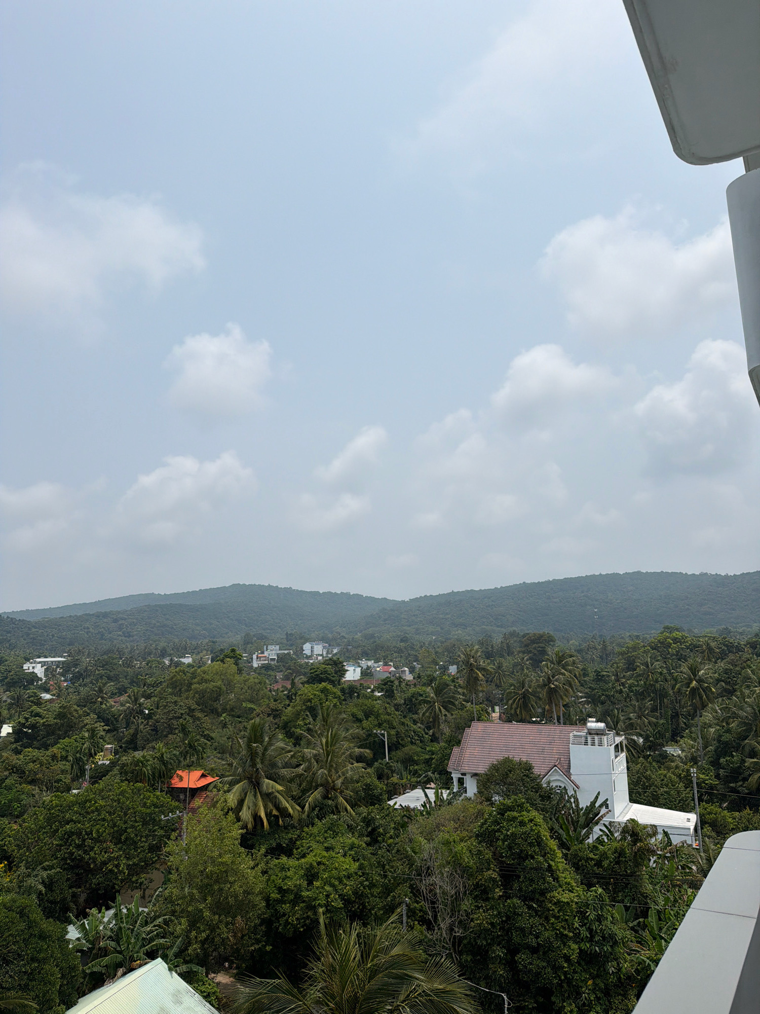 This is an aerial view of a densely vegetated landscape in Phú Quốc, Vietnam, on an overcast day. Lush green trees, including palm trees and banana trees, dominate the foreground and mid-ground, interspersed with several buildings.
One prominent building in the mid-ground is a white, multi-story structure with a tiled, reddish-brown roof and a white tower-like extension. This building appears to be a house or a small resort. Another building with a bright orange roof is visible further to the left, also nestled among the trees.
In the distance, a range of rolling green hills forms the backdrop. Scattered throughout the landscape are smaller white buildings, suggesting a small town or village. The sky is a pale blue with scattered white clouds. A portion of a light-colored balcony railing is visible in the upper right corner of the image, indicating the viewpoint is from a high elevation, likely a hotel or a residential building.
There are no people visible in the image. The scene is tranquil and dominated by nature and residential structures.