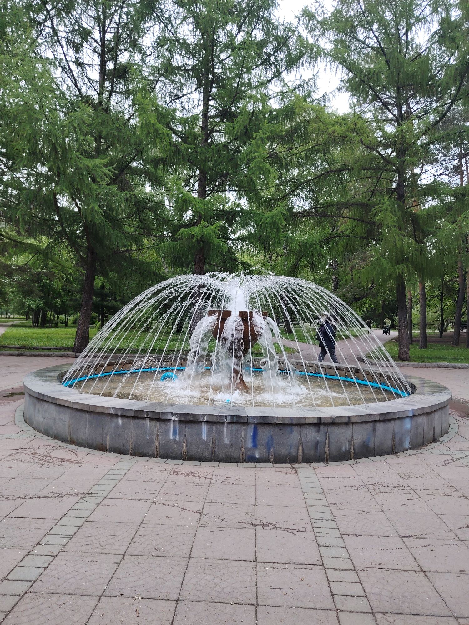 A full shot shows a fountain in a city park in Unknown, Unknown, with trees and people visible in the background. The fountain is round, with a gray outer edge and a pool of water at the center. The water is arched from the center into the pool. There is a person walking behind the fountain. The ground around the fountain is paved with square tiles. There are green trees behind the fountain.