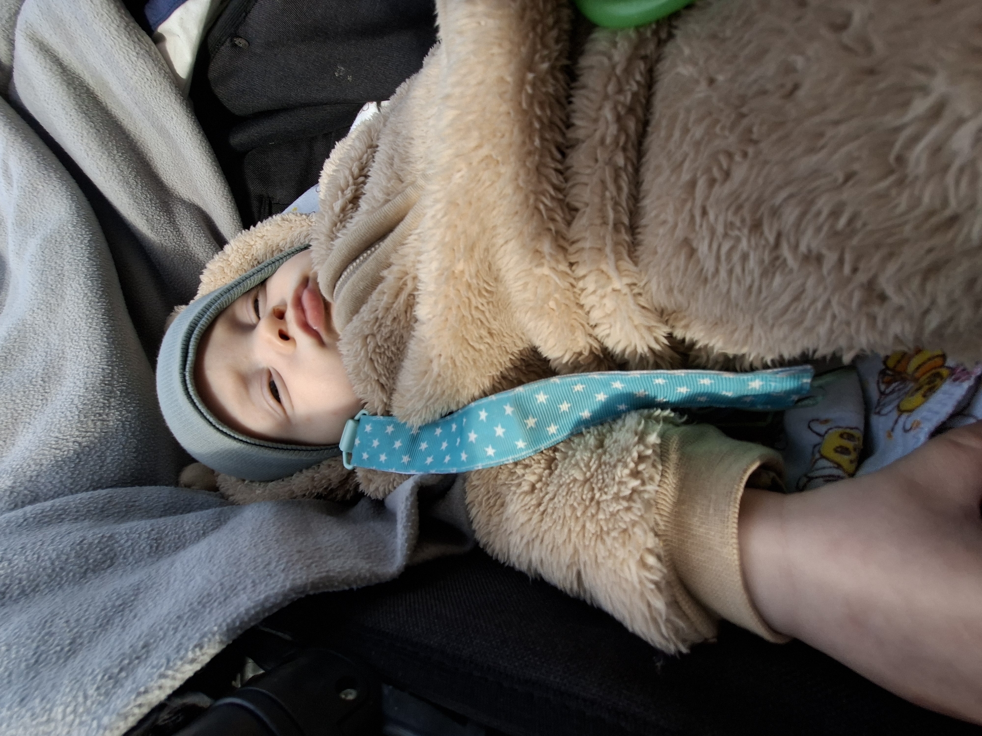 An infant is depicted resting in a black padded stroller or car seat, viewed from an overhead angle. The baby wears a light blue beanie and a fluffy, light brown, bear-themed full-body garment with ribbed tan cuffs. A light blue strap patterned with white stars is positioned across the baby's upper chest. The baby's eyes are mostly closed, and the mouth is slightly parted. A grey textured blanket covers the upper left portion of the frame and extends behind the baby's head. In the lower left, an adult's hand is visible near the baby's arm. Beneath the baby's left arm, a glimpse of patterned fabric with yellow cartoon bees is discernible. A green object is partially visible on the far right side of the frame. Natural light illuminates the scene, casting soft shadows. The location is in the city of Unknown, Unknown.
