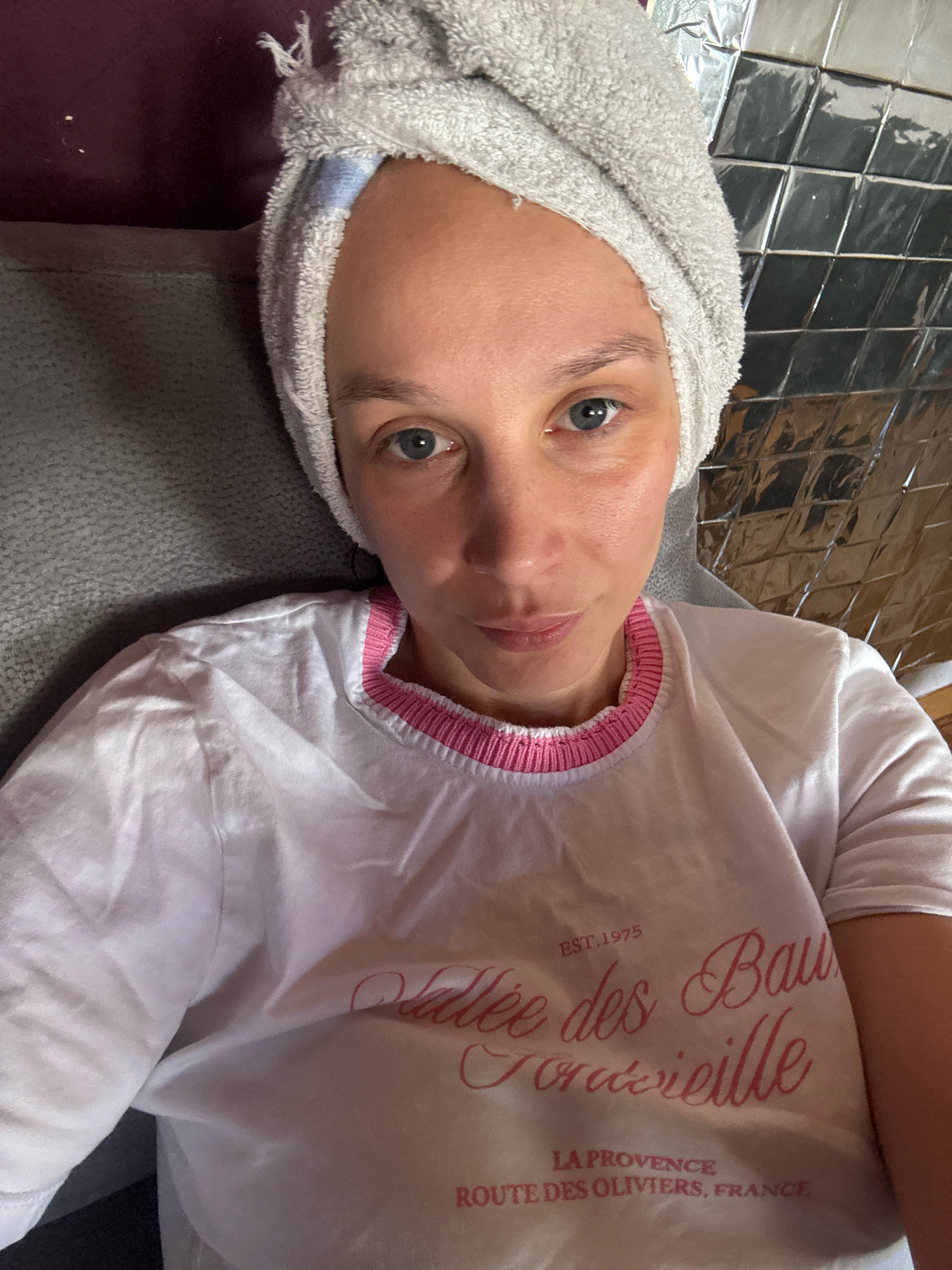 A woman with blue eyes and brown hair wrapped in a gray towel is taking a selfie. She is wearing a white t-shirt with a pink trim. The background consists of a textured gray surface and a reflective, tiled surface. The image does not provide any information to locate the scene in Überherrn, Germany.