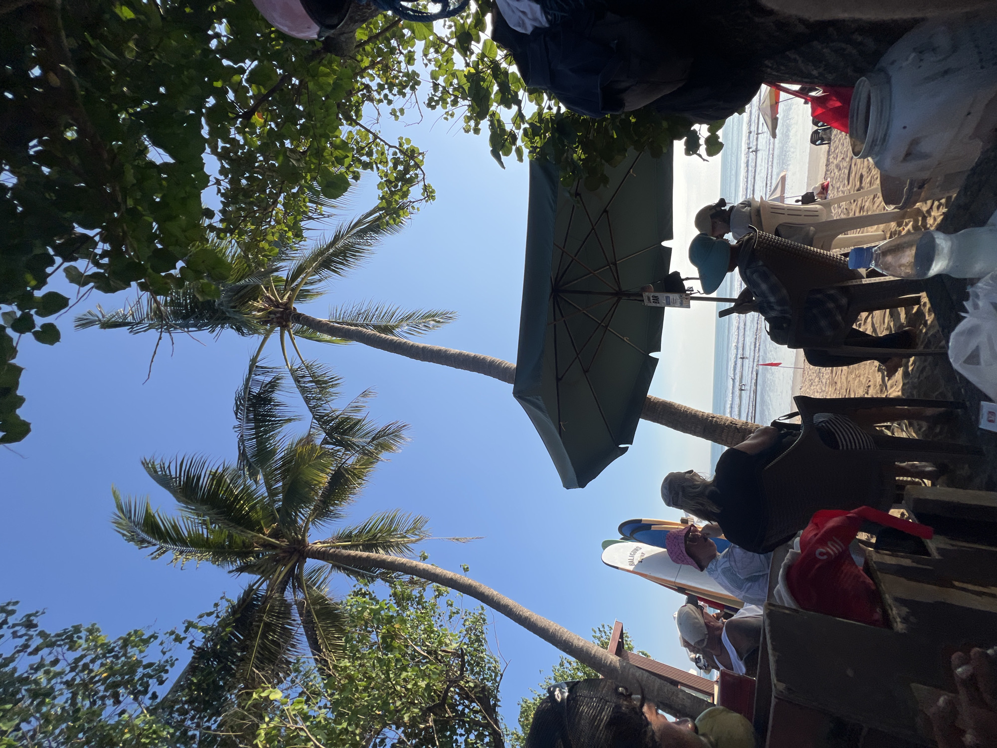 The image depicts a beach scene in Kuta, Indonesia, on a clear, sunny day. Two tall palm trees frame the view, with their fronds reaching towards the bright blue sky. Beneath the trees, a group of people are seated at tables on the sand, enjoying the beach atmosphere.
Several surfboards are propped up behind the seated individuals, suggesting a surf-friendly location. Some of the people are wearing hats, including a pink bucket hat and a light-colored baseball cap. One person is looking at their phone, while others appear to be conversing or relaxing. A large green umbrella provides shade for one of the seating areas.
In the background, the ocean stretches out to the horizon, with gentle waves breaking on the shore. A few people are visible in the water, some appearing to be surfing or swimming. Other beach umbrellas and chairs are scattered along the sandy expanse. The overall impression is one of a relaxed and idyllic beach setting, likely a popular spot for both locals and tourists in Kuta.