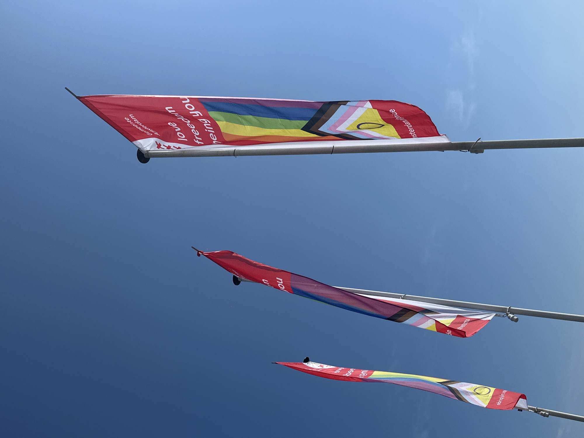 The image shows three pride flags flying against a clear blue sky. The flags are red with rainbow stripes and text. The text on the largest flag reads "Gemeente Amsterdam" and "love freedom being you". The flags are attached to poles. The image appears to have been taken in Amsterdam, Netherlands, as indicated by the text on the flag. There are no people visible in the image.