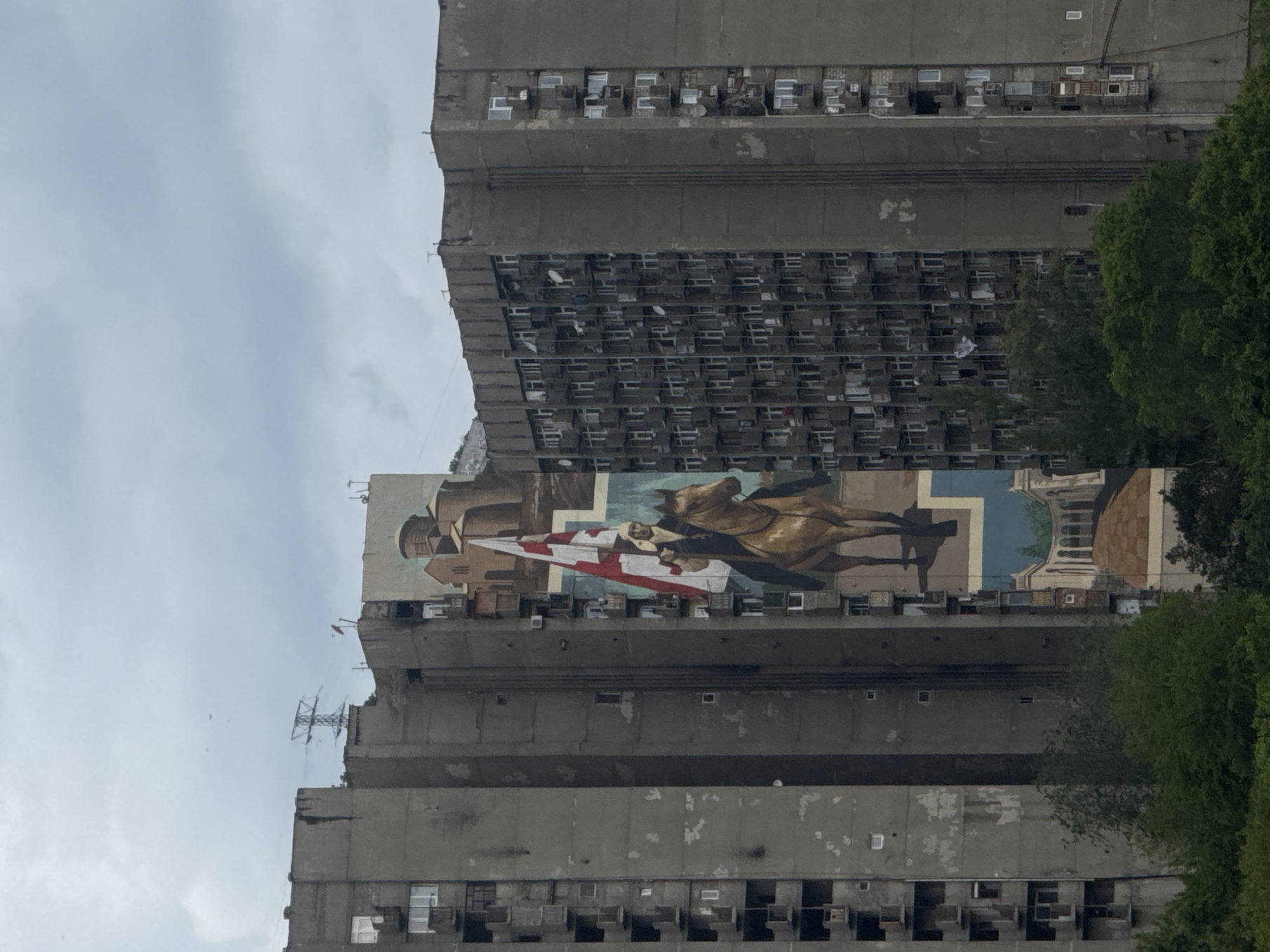 The image shows a tall, Soviet-era apartment building in Tbilisi, Georgia. A mural of Saint George slaying the dragon is painted on a large section of the building's façade. The building is concrete and grey, with many small windows. Some trees are visible on the right side of the frame. The sky is overcast. A communication tower is visible to the left of the building, on the upper midsection.