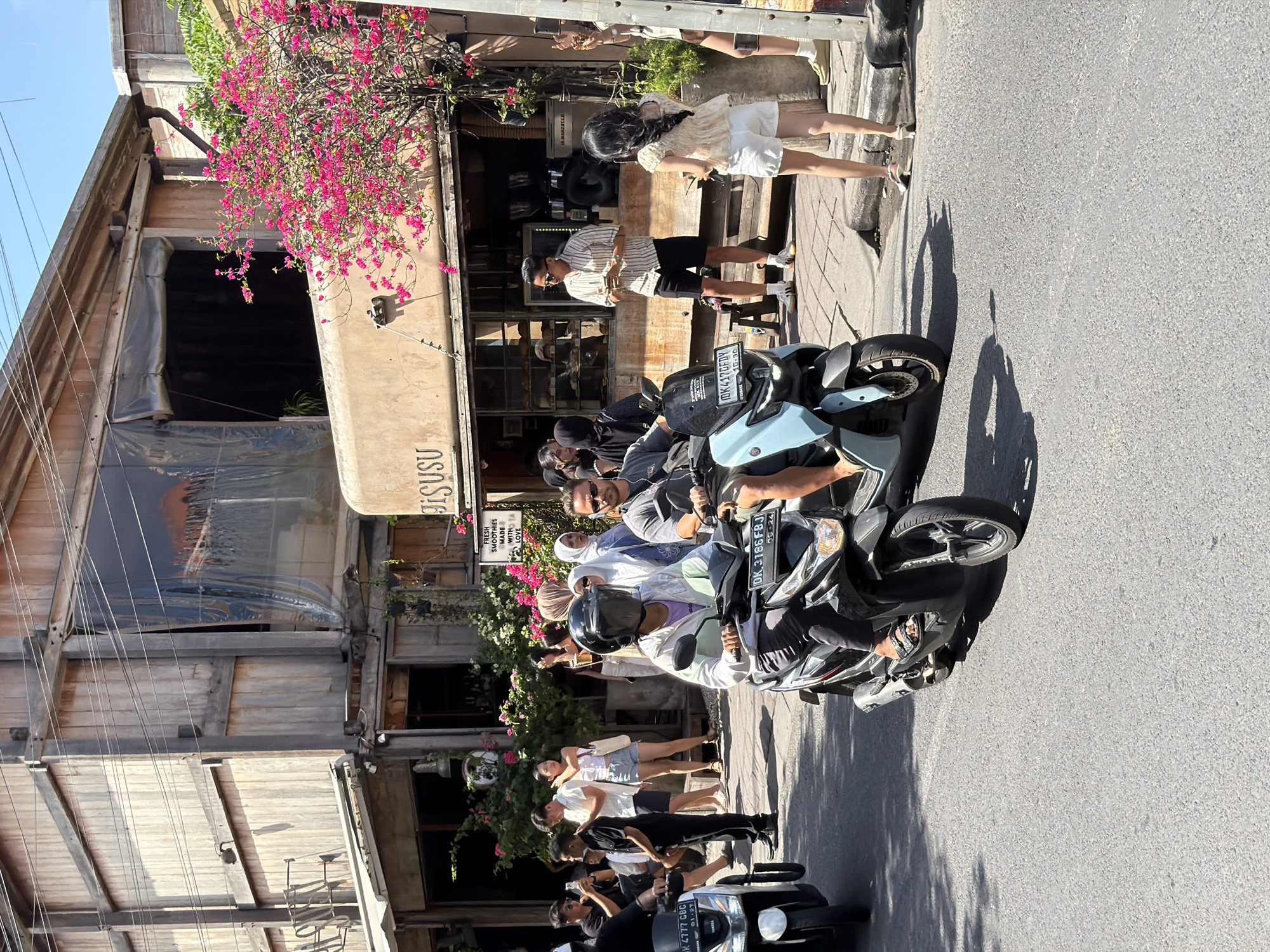 In North Kuta, Indonesia, on a sunlit day, a street scene unfolds with several people on or near motorcycles in front of a building. The building features a rustic wooden facade with large windows, and a sign reading "JISUSU". Pink bougainvillea flowers cascade from above the building. In the foreground, two individuals are riding scooters. The scooter on the left is black and silver, ridden by a person wearing a helmet with a dark visor and a light-colored jacket. The scooter on the right is light blue, ridden by a man in a gray t-shirt and sunglasses. Behind them, a group of people are gathered on the sidewalk. Some are taking photos with their phones. Others appear to be patrons of the establishment, with one man in a striped shirt and shorts holding a cup. A woman in a white top and shorts stands near the entrance. The street is paved with asphalt, and shadows from the motorcycles and people are cast across it. A white sign with black text is visible on the building, stating "FRESH SMOOTHIES MADE WITH LOVE".