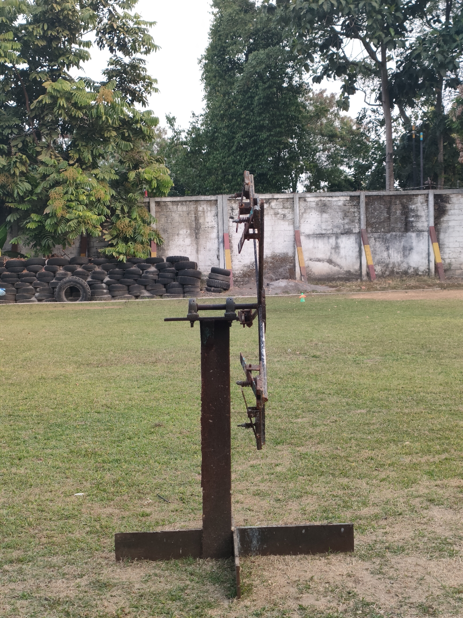 The image depicts an outdoor scene with a rusty metal apparatus in the foreground, standing on a grassy field. The apparatus appears to be an old, manual machine with various levers and wheels. In the background, there is a concrete wall that forms a boundary, with a pile of stacked tires behind it. Several trees are visible behind the wall, and the sky is overcast. The environment suggests a sports or training facility. The location is identified as Banguntapan, Indonesia, though no specific landmarks are visible to confirm this. There are no people or animals in the image.
