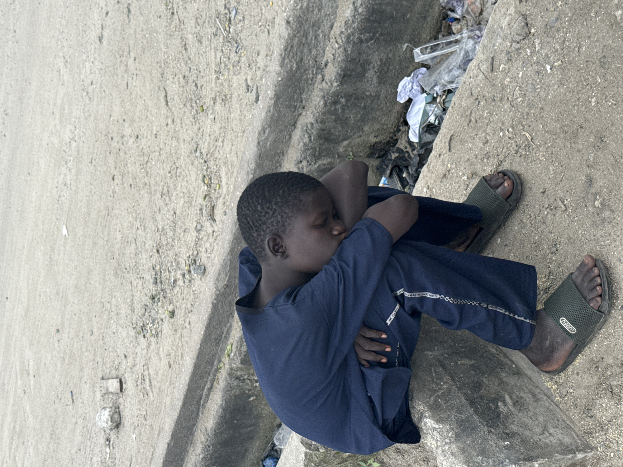 A young Black boy, wearing a dark blue long-sleeved shirt and dark pants with white trim, sits with his head bowed and resting on his arms, his eyes closed. He wears dark green slip-on sandals. The boy is seated on a concrete curb, with a dirt and debris-covered ground to his right and a concrete drainage ditch to his left. The area appears to be an outdoor urban setting in Satellite Town, Nigeria.