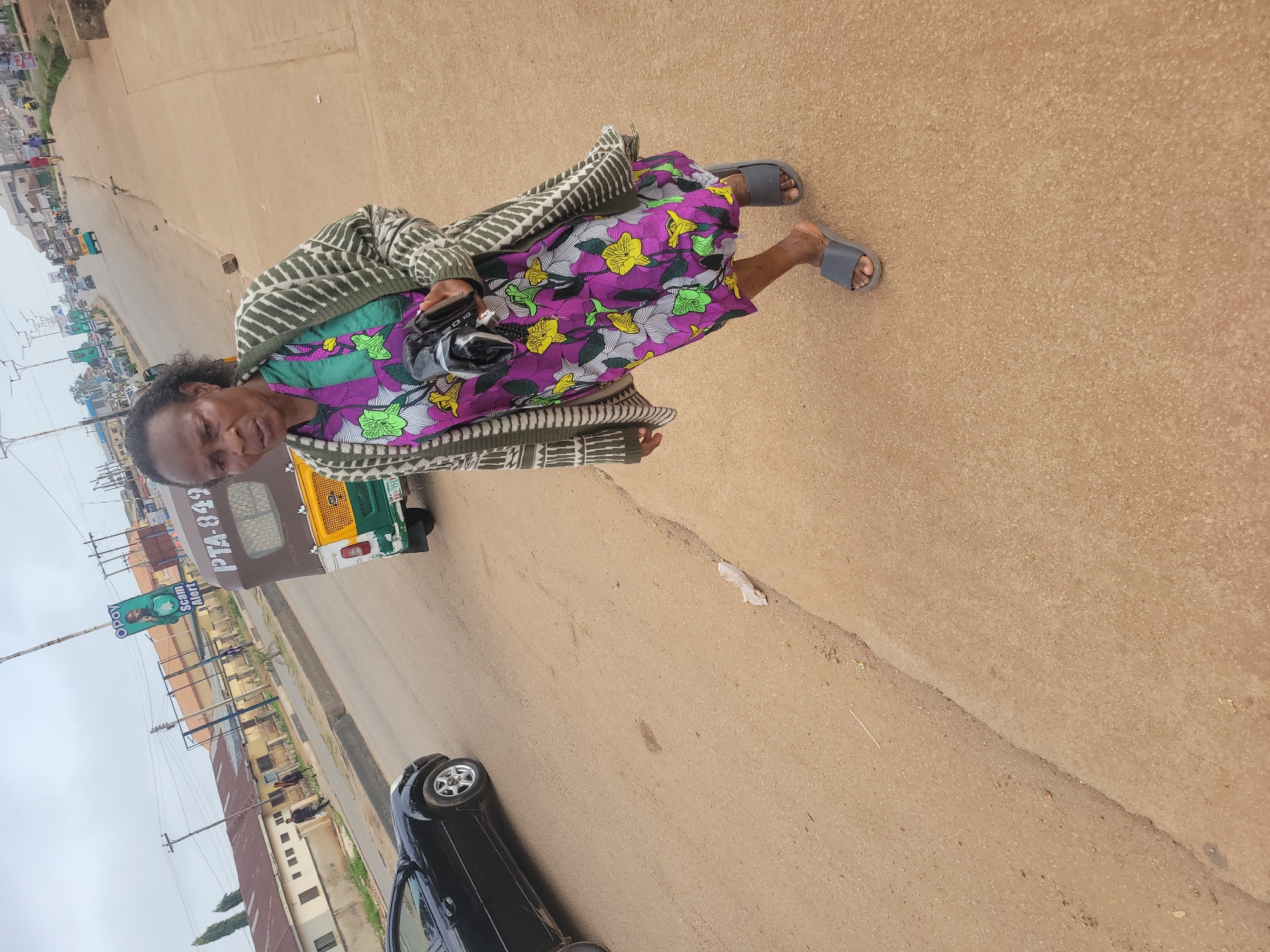 An elderly African woman is captured mid-stride on a paved road in Gura Top, Nigeria. She is wearing a vibrant purple and green floral dress and a patterned cardigan, with gray slide sandals on her feet. In her right hand, she holds a black object, possibly a camera. To her left, a black car is parked on the side of the road. In the background, a motorized tricycle with the license plate "PTA-849" is visible, along with other vehicles and buildings lining the street. The sky is overcast, suggesting a cloudy day. The road appears to be a main thoroughfare with moderate traffic.