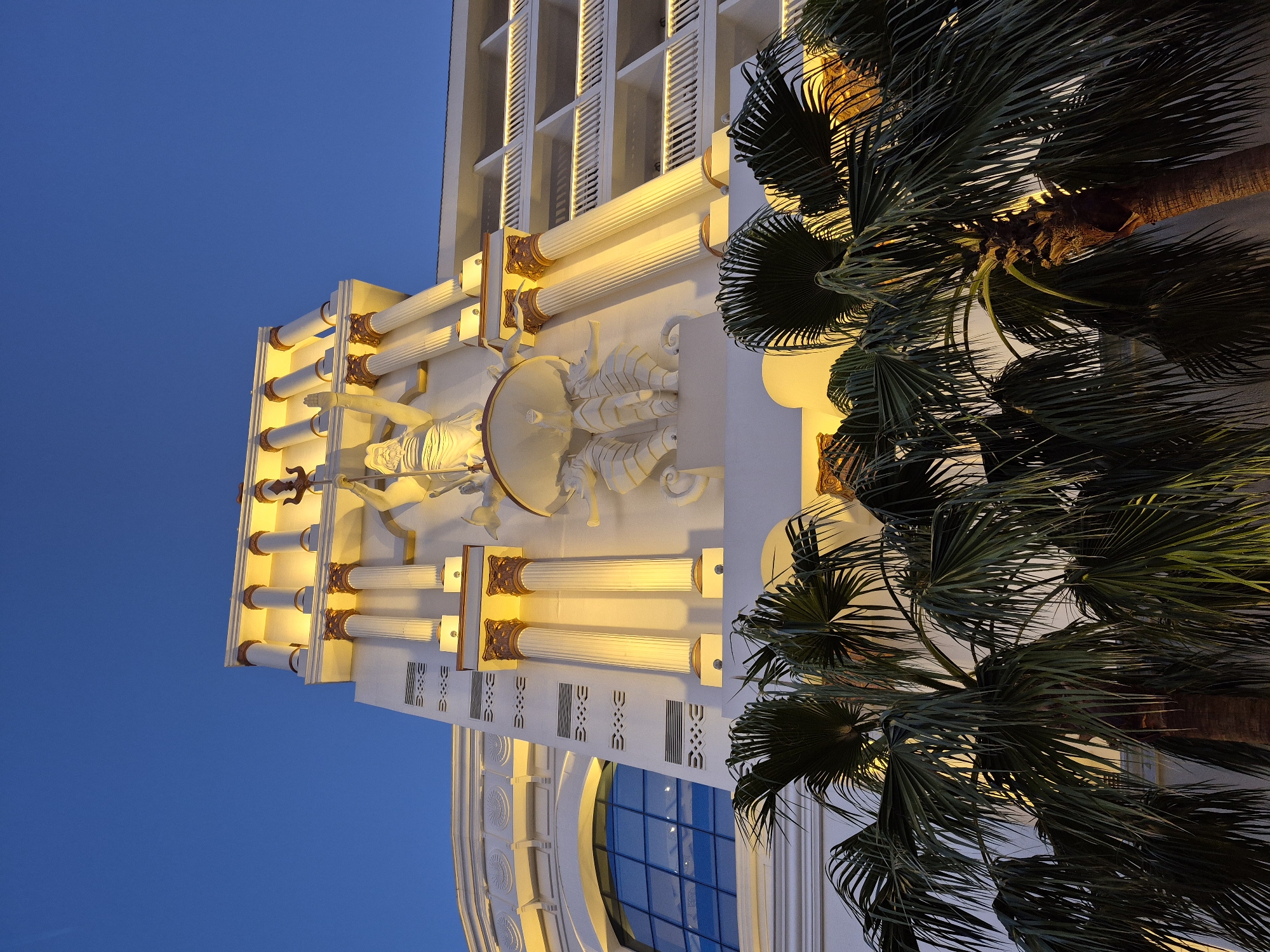 A low-angle shot depicts a building in Alanya, Turkey, adorned with classical architectural elements. The façade features illuminated Corinthian columns, intricate white sculptures of mythological figures and sea creatures, and decorative geometric patterns. A large window with blue panes is visible. A palm tree with dense foliage frames the right side of the building. The sky is a gradient of blue, suggesting twilight. The image showcases the architectural style and landscaping of a building in Alanya.