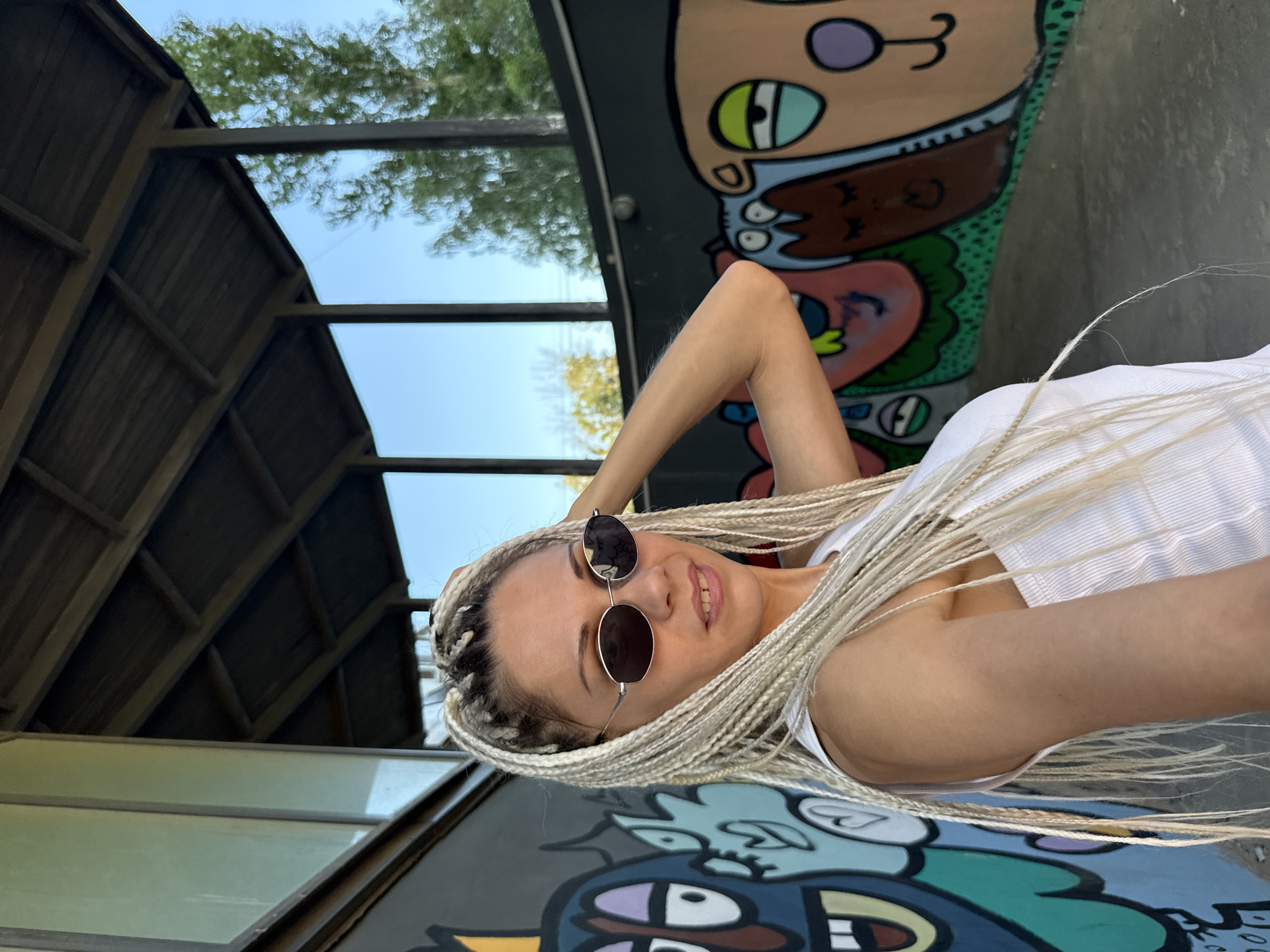 A young woman with blonde braids and sunglasses is standing in front of a graffiti-covered wall. She is wearing a white tank top. The background shows a roof structure and trees against a clear blue sky. This scene is in the city of Korobovo, Russia.