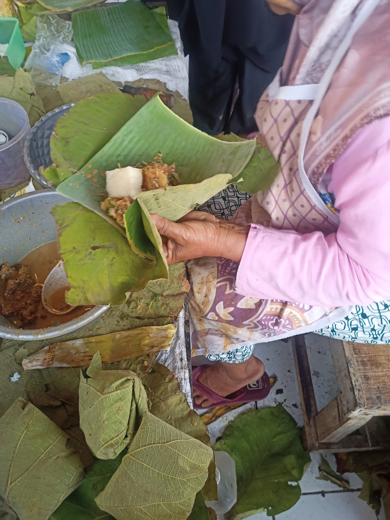 This image captures a street food vendor in Nganti, Indonesia, preparing a meal. The vendor, wearing a pink long-sleeved shirt and a patterned apron, is seen wrapping food in large green leaves. A metal bowl containing a savory dish, likely a stew or curry, is visible, along with a ladle. Several portions of food are already wrapped in leaves and arranged around the workspace, some of which are resting on a larger, dried leaf. The scene appears to be set up on a wooden surface, with other food preparation materials and a plastic cup also present. The overall impression is of a humble, outdoor food stall where traditional methods are used for serving meals.
