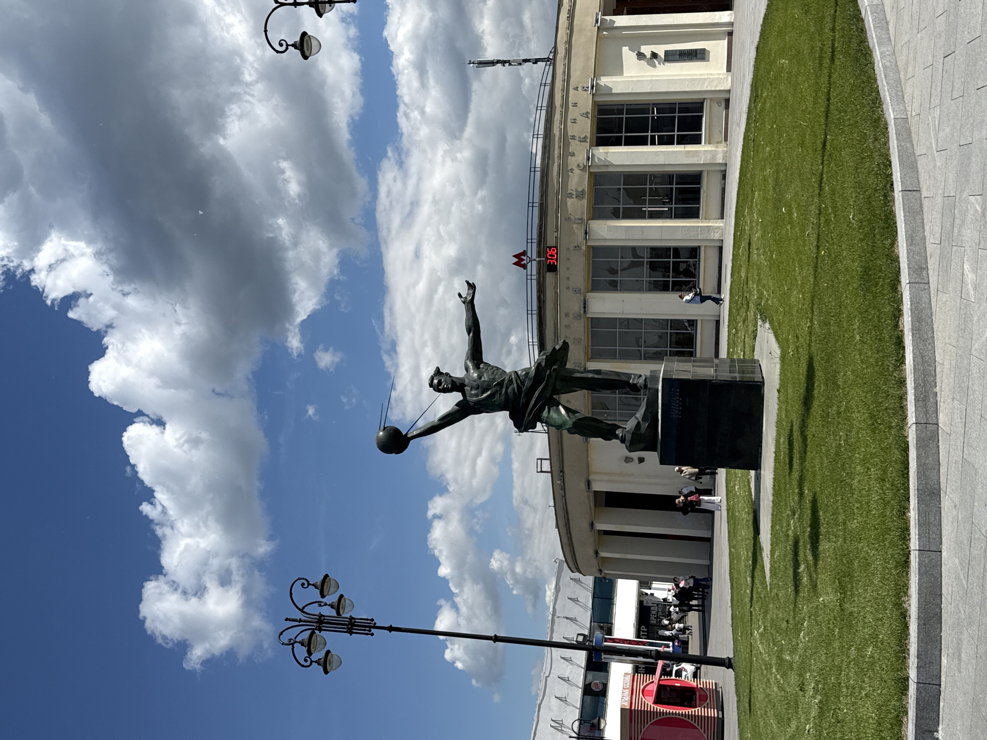 The image captures an outdoor urban scene in Moscow, Russia, featuring the "Conquerors of Space" monument at VDNKh. A large bronze statue of a male figure, depicted in dynamic motion, dominates the foreground. He is shown holding aloft a sphere with antennae, emblematic of the Sputnik satellite, and stands on a dark, rectangular pedestal.
In the background, a grand, light-colored building with large windows serves as the VDNKh Metro station entrance. Prominently displayed on its facade are the red "METPO" (Metro) logo and a digital clock showing "3:06", suggesting the time is 3:06 PM. Above the windows, the Cyrillic text "ВЫСТАВКА ДОСТИЖЕНИЙ НАРОДНОГО ХОЗЯЙСТВА" (Exhibition of Achievements of National Economy) is inscribed.
The setting includes neat green lawns and light-colored paved walkways. Several pedestrians are visible in the background, walking near the building and on the paved areas. Two ornate street lights with multiple lamps are also present in the scene. A red kiosk-like structure with partially visible Cyrillic text, including "ЦЕНТР" (Center) and "АРЕНДА" (Rental), can be seen on the right side.
The weather is bright and sunny, with a clear blue sky interspersed with white and grey clouds, indicating a pleasant day. Shadows are cast on the grassy areas. The overall impression is one of a significant public space, bustling gently with activity.