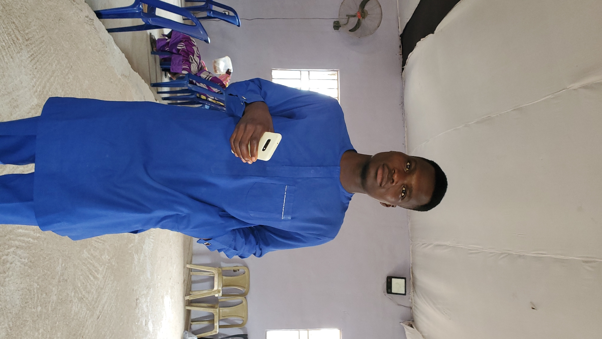 A young man wearing a blue traditional outfit stands in a room with a white ceiling. He is holding a white mobile phone in his right hand. The man is looking towards the camera with a neutral expression. In the background, there are several blue plastic chairs and some beige chairs arranged in rows. A woman in a purple and patterned outfit is seated in one of the blue chairs. There is a fan mounted on the wall to the left of the man. The room appears to be sparsely furnished and has a simple concrete floor. The setting suggests a community gathering or a place of worship. The image does not provide specific clues to pinpoint the exact location within Jalingo, Nigeria.