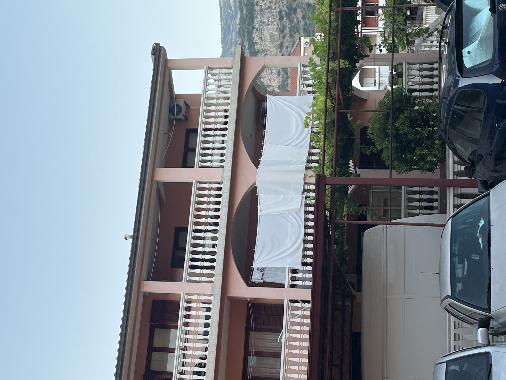 The image captures a section of a multi-story residential building in Budva, Montenegro. The building has a pink facade and features multiple balconies with white balustrades. A large white sheet is hung across one of the lower balconies, suggesting laundry is being dried. In front of the building, a white van is parked, partially obscuring the ground floor. To the left of the van, the silver roof and windshield of a car are visible, and to the right, the dark blue rear of another car. Lush green foliage, likely a grapevine canopy, is spread over a metal frame in the foreground, creating a shaded area. In the background, a rugged, mountainous landscape is visible under a clear, pale blue sky. The architectural style suggests a typical Mediterranean dwelling, common in coastal towns like Budva.