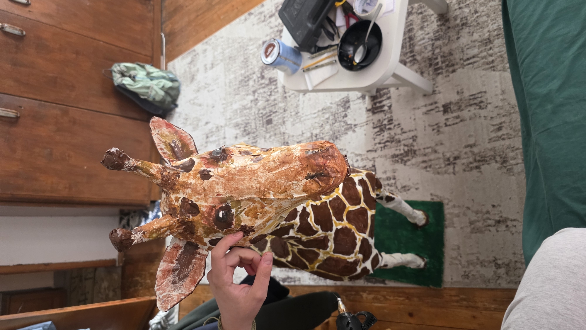 A person holds a papier-mâché giraffe in a room with a dresser and a white table. The giraffe has a brown and white head with brown spots, and a brown body with white legs. The person's hand is visible near the giraffe's ear. The dresser has wooden drawers. The white table has a black bowl, a blue container, and other items on it. The floor is covered in a patterned rug. There is no indication of the location being in Blagoevgrad, Bulgaria.