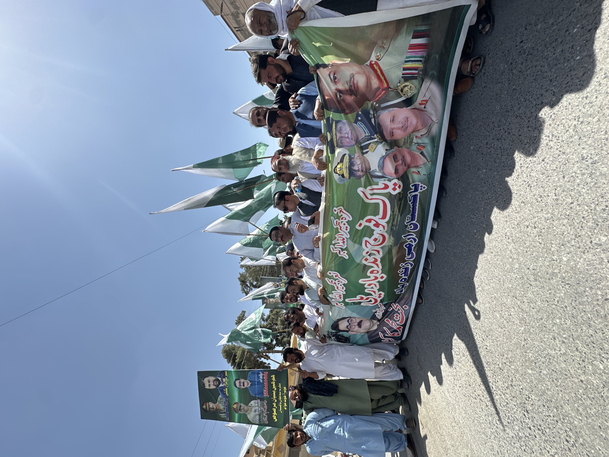 A group of men in Quetta City Tehsil, Pakistan, are marching down a road. They are holding Pakistani flags and banners. One banner features portraits of various Pakistani military figures and is inscribed with the words "Pakistan Army Zindabad." Another sign shows two men in suits. The setting is outdoors on a sunny day.