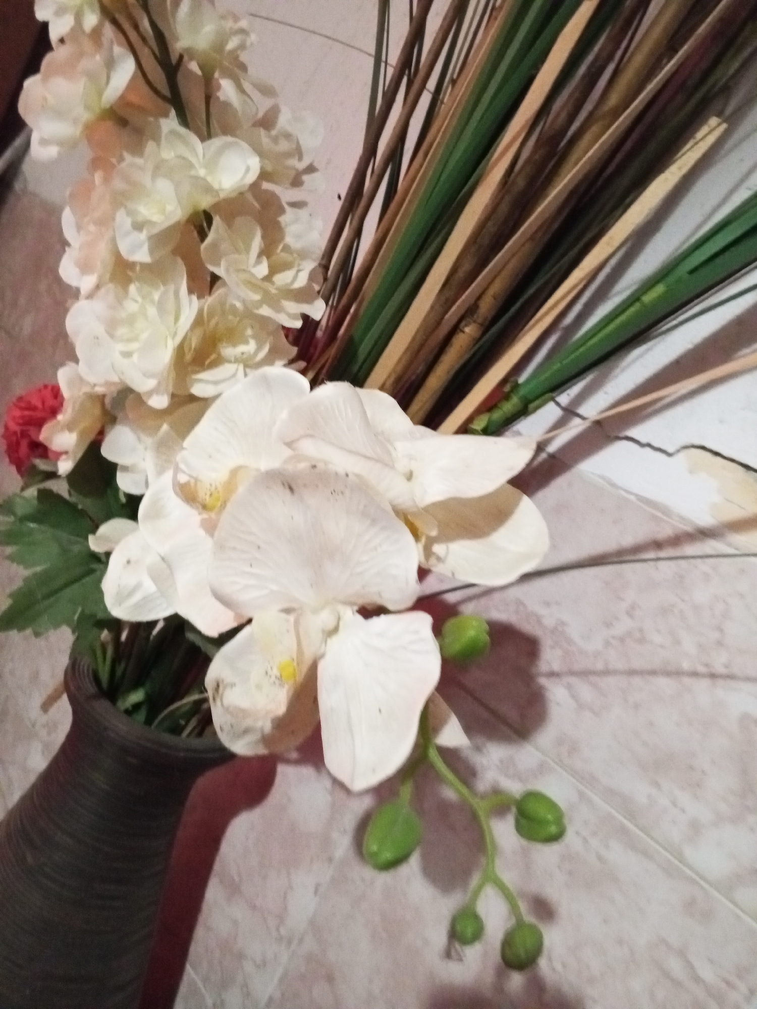 A still life depicts an arrangement of artificial flowers and reeds in a textured, brown vase. The flowers are primarily white, with a cascading effect. A small red flower is also visible. The reeds are a mix of brown and green. The arrangement sits on a surface with a tiled pattern. The location is Unknown, Unknown.