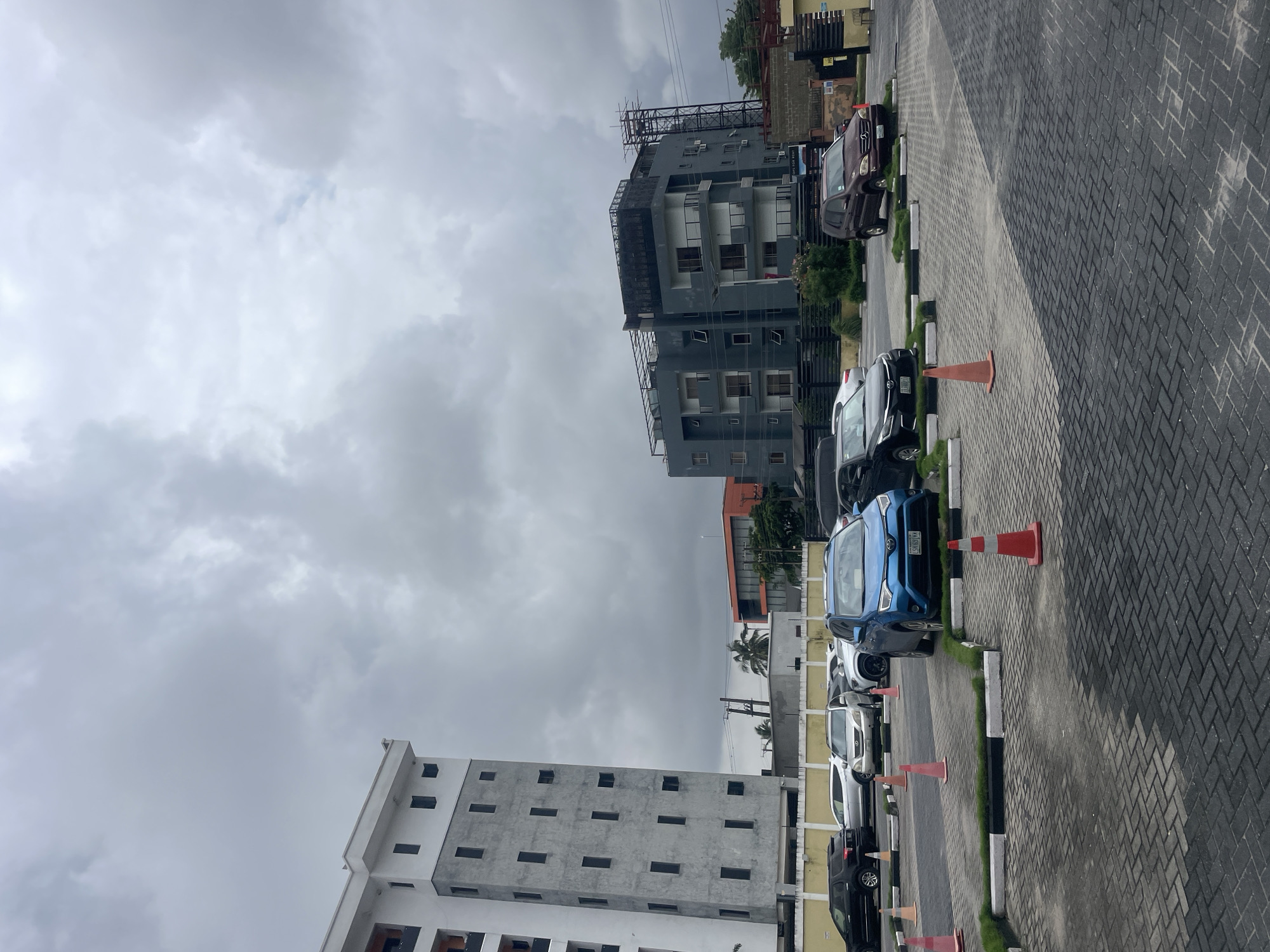 The image depicts an outdoor scene in the city of Moba, Nigeria, under an overcast sky. In the foreground, a paved parking lot is visible, laid with interlocking bricks in a pattern of light and dark grey. Several cars are parked in designated spots marked by white and black curbs and patches of grass. Traffic cones are scattered throughout the parking area.
Dominating the left side of the image is a tall, modern building with a white facade and gray concrete sections, featuring numerous rectangular windows. To the right of this building, another contemporary structure is visible, painted in dark gray with white accents and multiple balconies. Further in the background, a smaller orange building can be seen, along with trees and other structures. The general environment suggests an urban or semi-urban setting, possibly a commercial or residential complex. No people are visibly present in the scene.