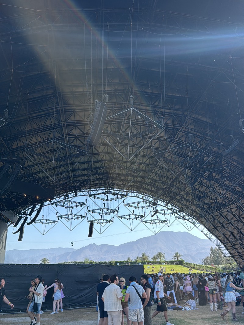 The image shows a crowd of people gathered at a large outdoor venue in Palm Desert, United States. Above the crowd is a large, arched metal structure supporting rigging and speakers. Mountains and palm trees are visible in the background through the arch. People are standing, walking, and sitting on the ground. Some are conversing in small groups.