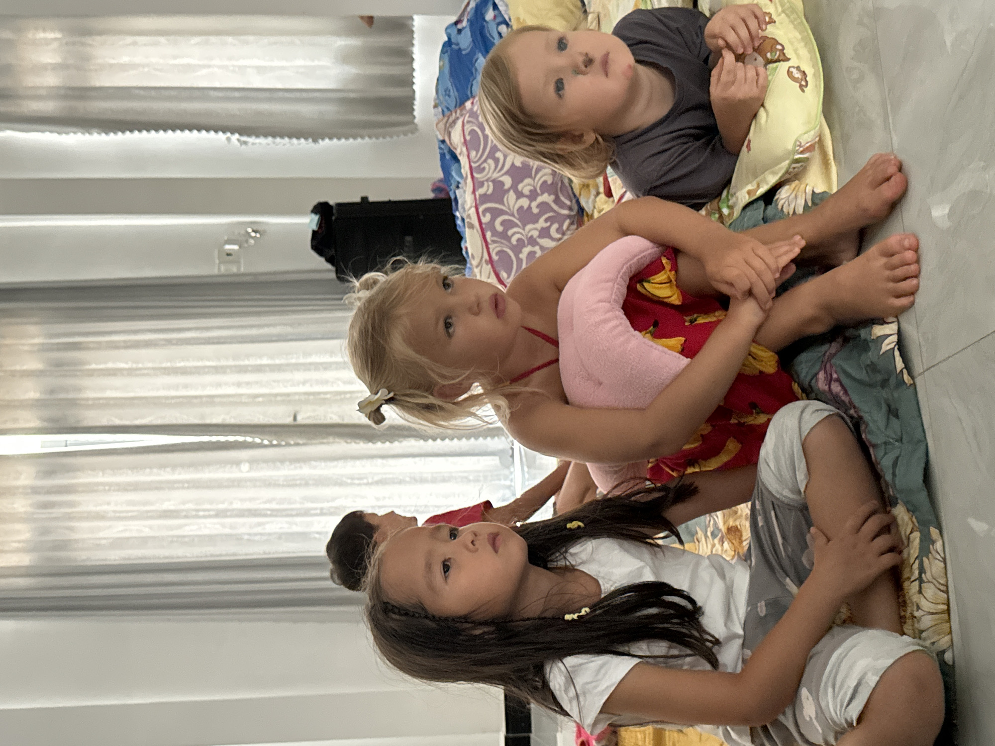 Three children are sitting on a bed or mattress on the floor in a room with white blinds. A young Asian girl with long dark hair and small yellow clips in her hair sits in the foreground, wearing a white t-shirt and gray patterned pants. Behind her is a slightly older blonde girl wearing a red dress with a pink plush object in her arms. Further back is a younger blonde child wearing a dark gray shirt. There is a dark object that could be luggage in the background near the blinds. The location is Mũi Né, Vietnam.