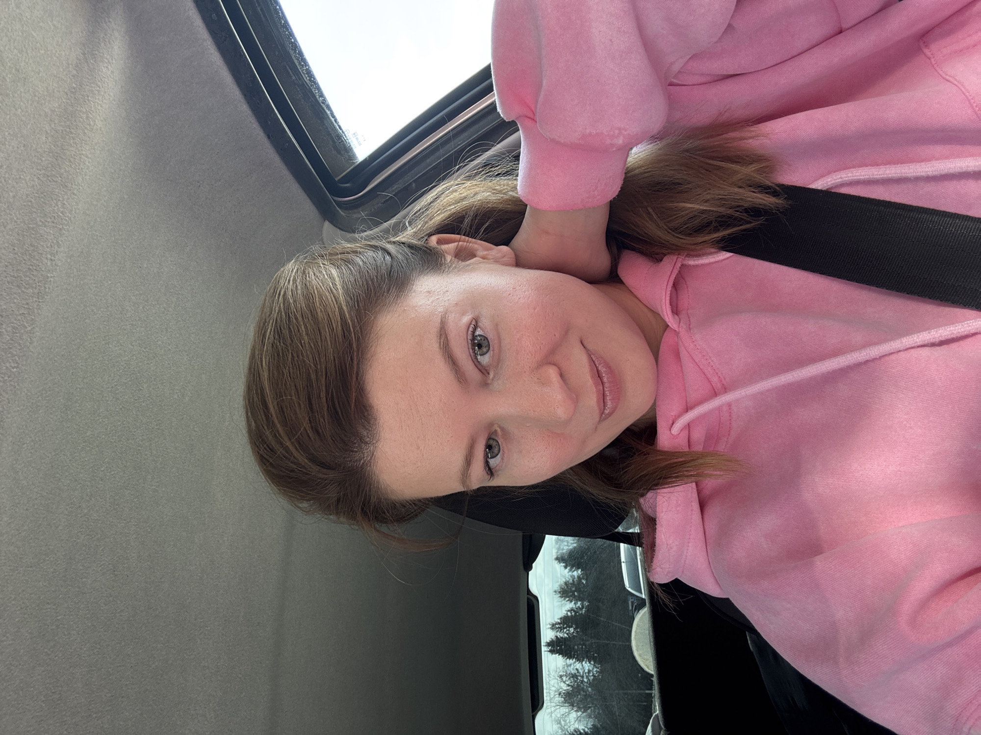 A woman with fair skin and light brown hair is sitting in a car, wearing a pink hooded sweatshirt and a black seatbelt. The interior of the car is gray, with a sunroof visible. Outside the car window, trees can be seen, suggesting a wooded area. Based on the information provided, the location could be Chaikovsky, Russia.