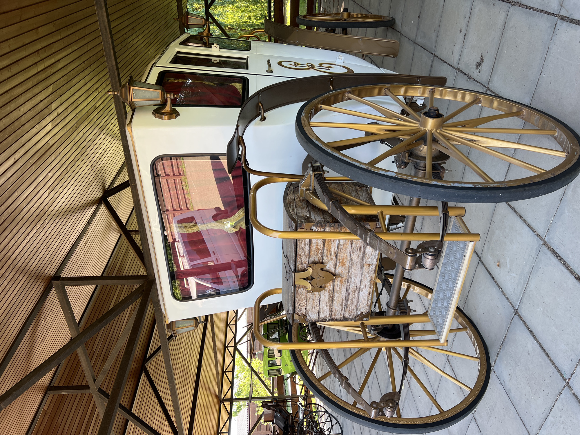An antique white carriage with gold trim and spoked wheels is parked under a wooden roof in the city of Plastunivska, Russia. The carriage has a wooden chest on its rear, a step for passengers, and two lamps on its exterior. The ground is paved with square tiles. In the background, other carriages and trees are visible.