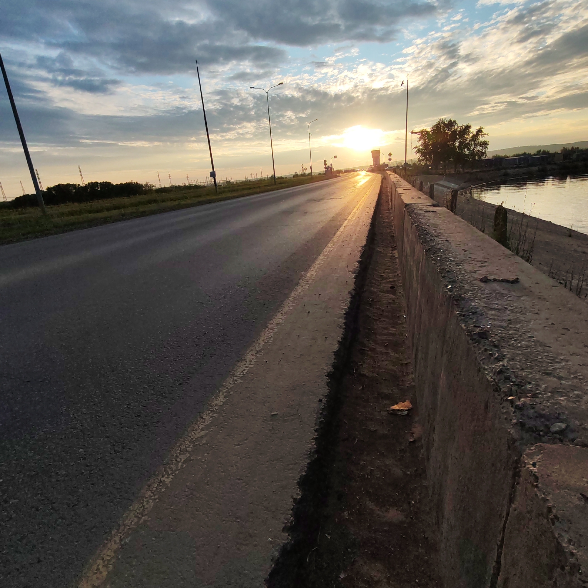The image shows a road with a concrete barrier on the right side, bordering a body of water. The sun is setting, casting a warm glow on the scene and reflecting off the road and water. The sky is filled with clouds, some of which are illuminated by the setting sun. There are streetlights along the road, and a few trees are visible in the distance. The location is on the embankment of the Kama River in Chaikovsky, Russia, near the hydroelectric power station. There are no people or specific actions depicted in the image.