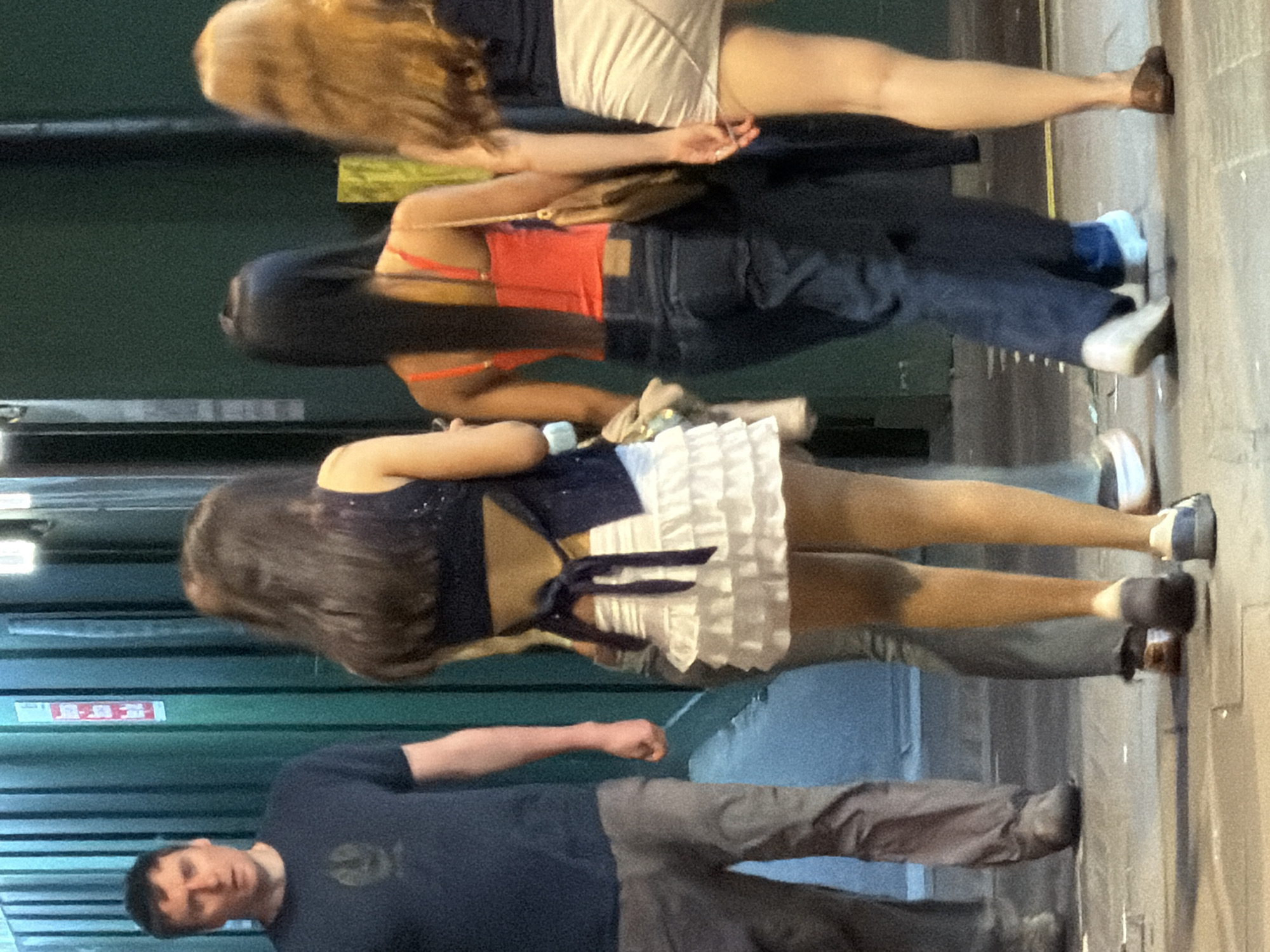A street scene shows four people. The person at the bottom is a male wearing a dark t-shirt and gray pants. Above him is a female wearing a navy and white short dress with black flats. The next person is a female wearing a red top and blue jeans, and white shoes. The person at the top is a female wearing a dark top and light shorts with brown flats. They are standing against a green wall on a city sidewalk. The location appears to be London, United Kingdom due to the architectural style and the type of bus visible in the background.