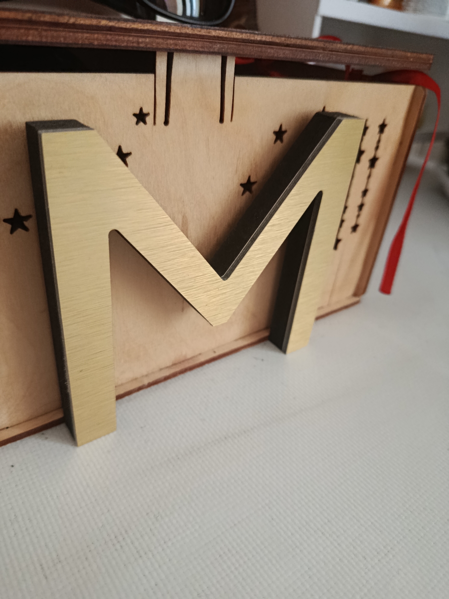 A close-up, eye-level, indoor shot focuses on a large, golden letter "M" with a dark edge, positioned diagonally on a light-colored wooden surface. This surface is part of a box-like structure with a dark wooden frame and red ribbon trailing from its side. The wooden surface is decorated with small, black star cutouts and vertical slots. The letter "M" is the most prominent object, appearing to be a free-standing decoration. The background is a plain, light-colored surface, possibly a table or tablecloth, with a subtle textured pattern. The lighting is soft and even, casting minimal shadows. The image is sharp, with a shallow depth of field that blurs the background. There are no people present in the scene. The scene is not indicative of any specific urban environment; it appears to be a still life arrangement.