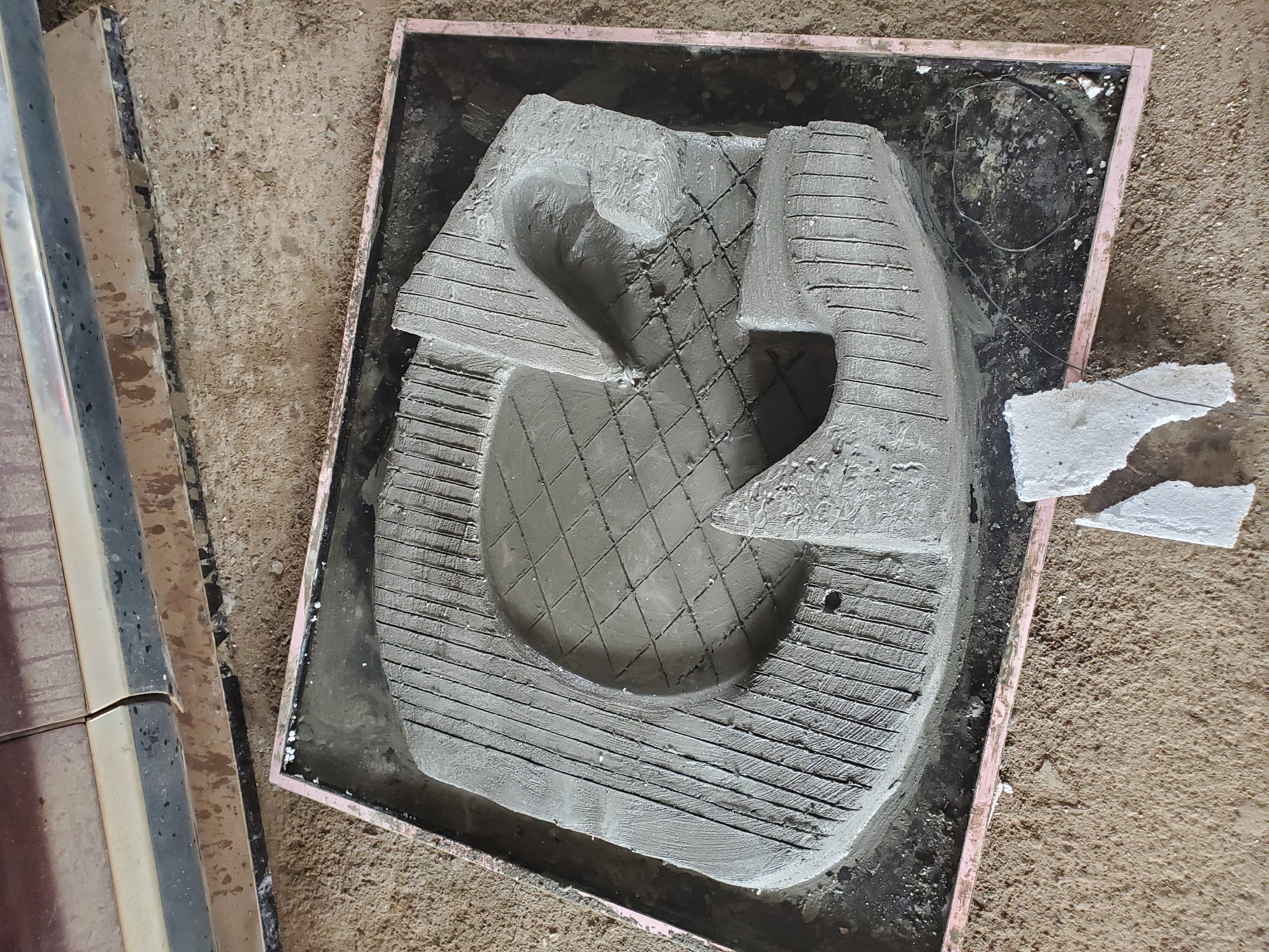 A concrete mold in the shape of the letter "G" sits in a pink rectangular frame on a dirt surface. The letter is partially filled with a wet, grey concrete mixture, which has been scored with a pattern of parallel lines and cross-hatching. To the right of the mold, the concrete forms a raised, curved shape with a rough texture. A small piece of white material, possibly Styrofoam, lies on the dirt in front of the mold, with a thin wire extending from it. In the background, the lower portion of a dark-colored vehicle is visible, its paintwork appearing dirty and streaked. The ground beneath and around the mold is composed of loose dirt and gravel. The scene is likely an outdoor area in Jos, Nigeria, where construction or crafting activities involving concrete are taking place.