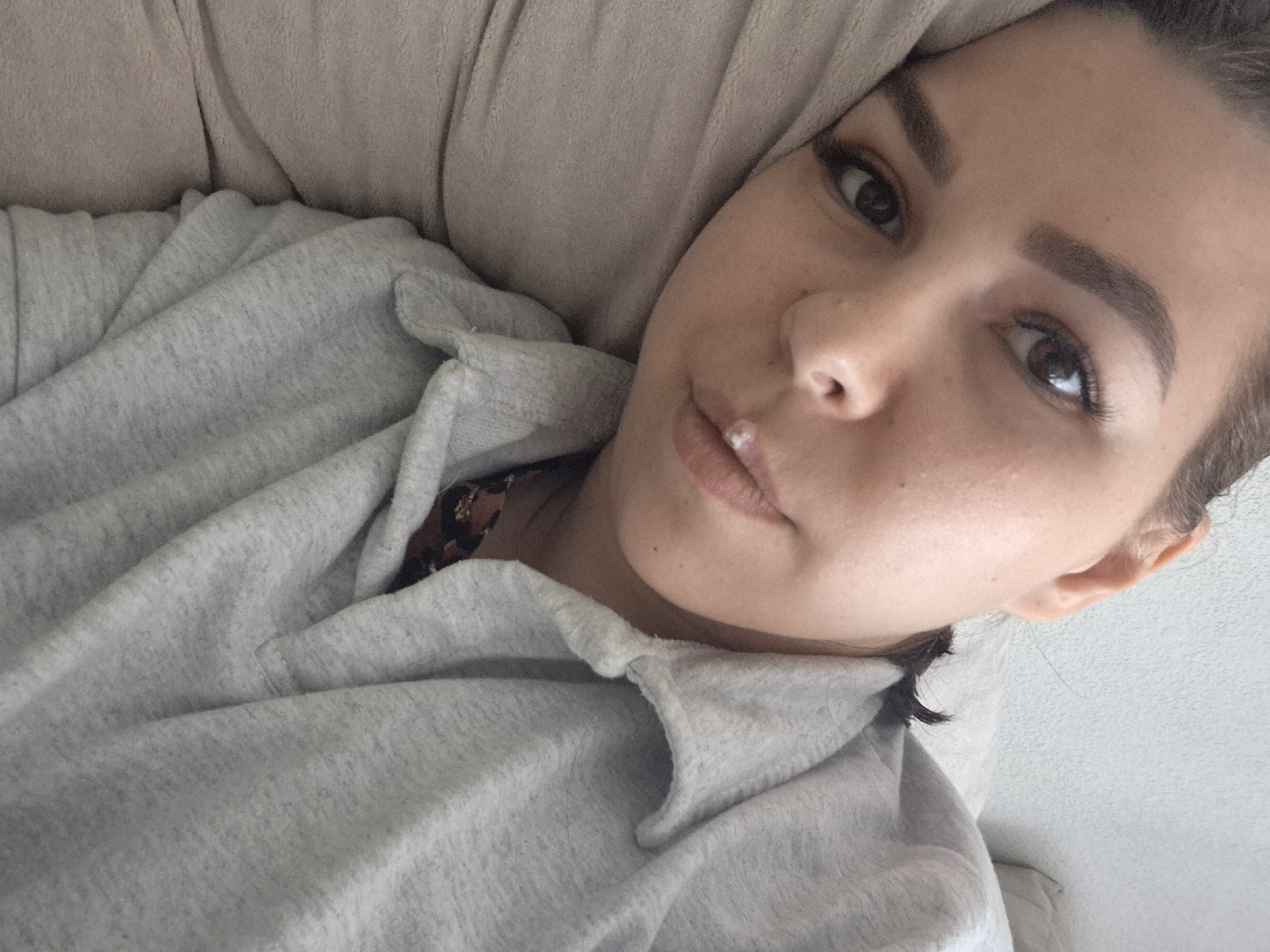 A woman with brown hair is lying down in a gray sweatshirt with a brown and black patterned shirt underneath. She is looking at the camera. The background is a beige color. There is no evidence to suggest the location is Tobolsk, Russia.