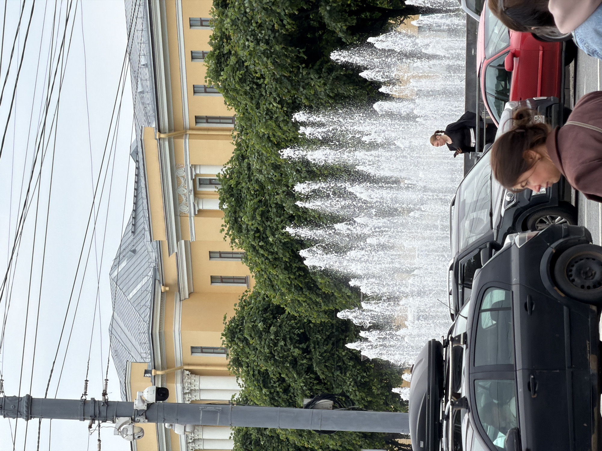 The image shows a street scene in Saint Petersburg, Russia, with a large yellow building in the background, partially obscured by trees and a fountain. The building has a grey, multi-pitched roof and classical architectural elements like columns and decorative friezes. In the foreground, several cars are parked or driving on the street. A black hatchback with a roof box is prominently featured on the left. To its right, a dark SUV and a red sedan are visible. A woman in a black outfit is standing near the fountain, looking at her phone. Another woman, facing away from the camera, is in the foreground, closer to the viewer. The sky is overcast. A tall lamppost with security cameras is on the left side of the image, along with overhead wires. The fountain sprays water high into the air, creating a misty effect.