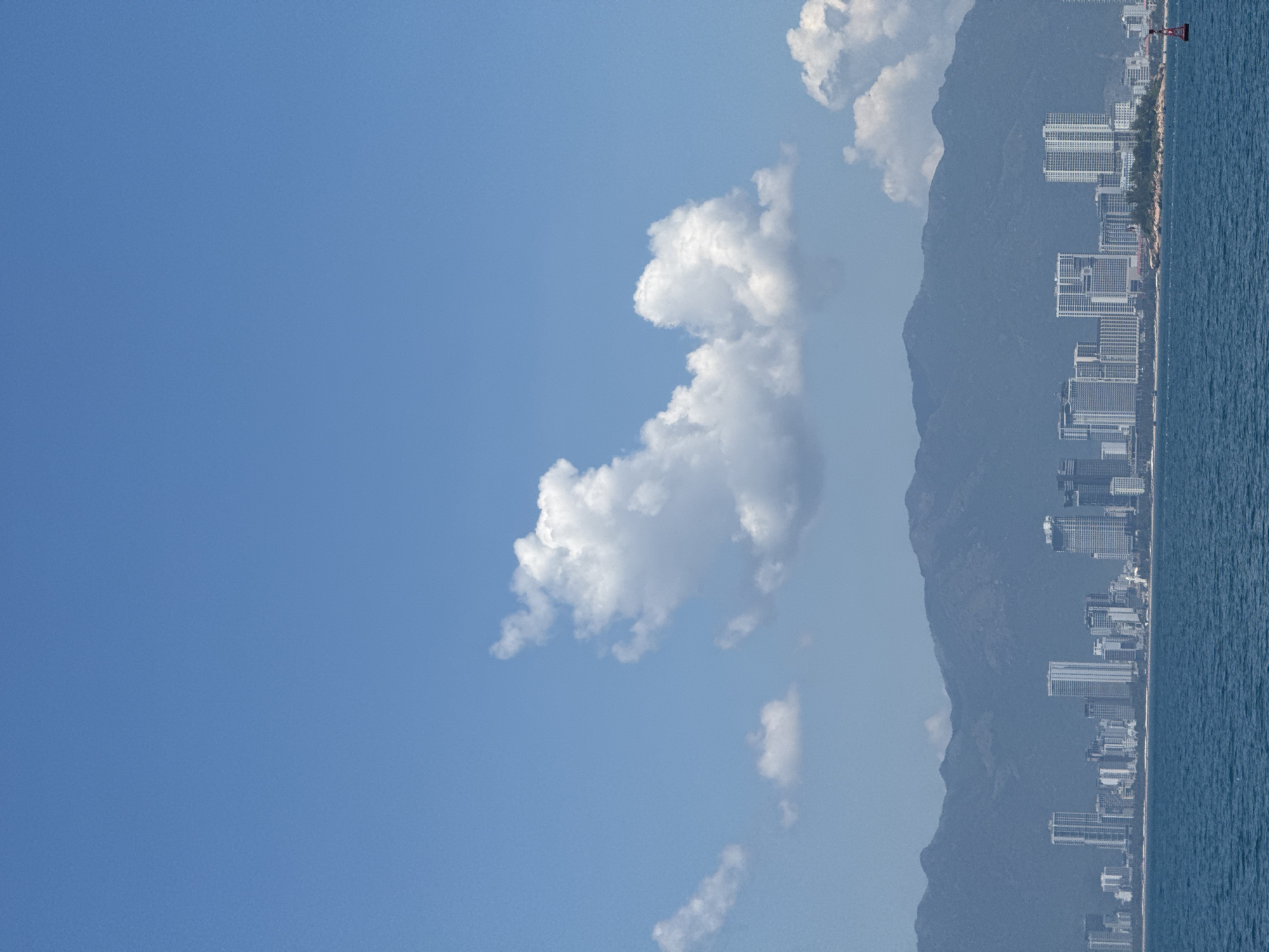 The image captures a panoramic view of a coastal city in Bac Nha Trang Ward, Vietnam, under a bright, clear blue sky dotted with fluffy white clouds. In the foreground, the deep blue ocean stretches out, with a visible wave pattern. The middle ground features a densely packed city skyline composed of numerous tall, modern buildings, including skyscrapers, apartments, and hotels. The architecture is predominantly white and gray, with varied window designs. Behind the city, a range of majestic, hazy blue mountains forms a dramatic backdrop. The scene appears to be taken from the water, possibly from a boat or the shore, looking towards the land. A red and white buoy is visible in the water to the right of the frame. There are no people or visible actions depicted in the image.