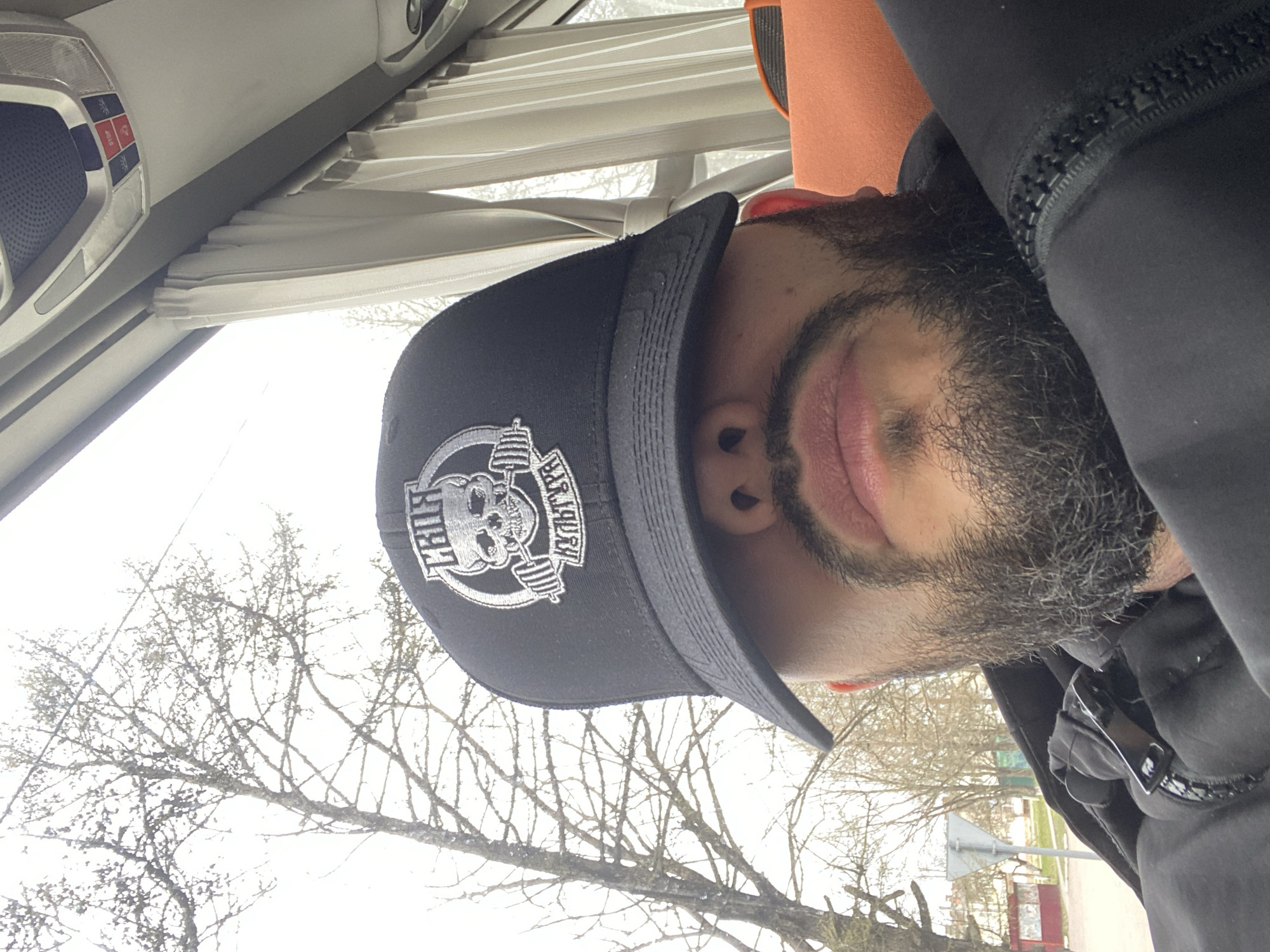 A close-up selfie captures a man inside a vehicle, looking towards the camera. The image is rotated clockwise. The man has a beard and mustache and is wearing a black baseball cap with a prominent white embroidered logo depicting a skull with a barbell and the text "PIRATE GYM" around it. He is also wearing a black hooded garment, possibly a jacket or hoodie, with a zippered detail visible near his neck where a pair of sunglasses is tucked in.
The setting appears to be the interior of a vehicle, likely a truck or bus cabin, given the overhead console with multiple buttons, including a red "STOP" button, and pleated window shades. Through the vehicle's window, bare trees are visible against a bright, overcast sky, suggesting a cold season like late autumn or winter. In the distant background, a road sign and some faint structures can be seen, hinting at an outdoor, possibly semi-rural or industrial, environment. The man's lips are slightly parted, but no specific emotion is clearly discernible. The overall lighting suggests it is daytime. The location context provided is Opolie, Russia.