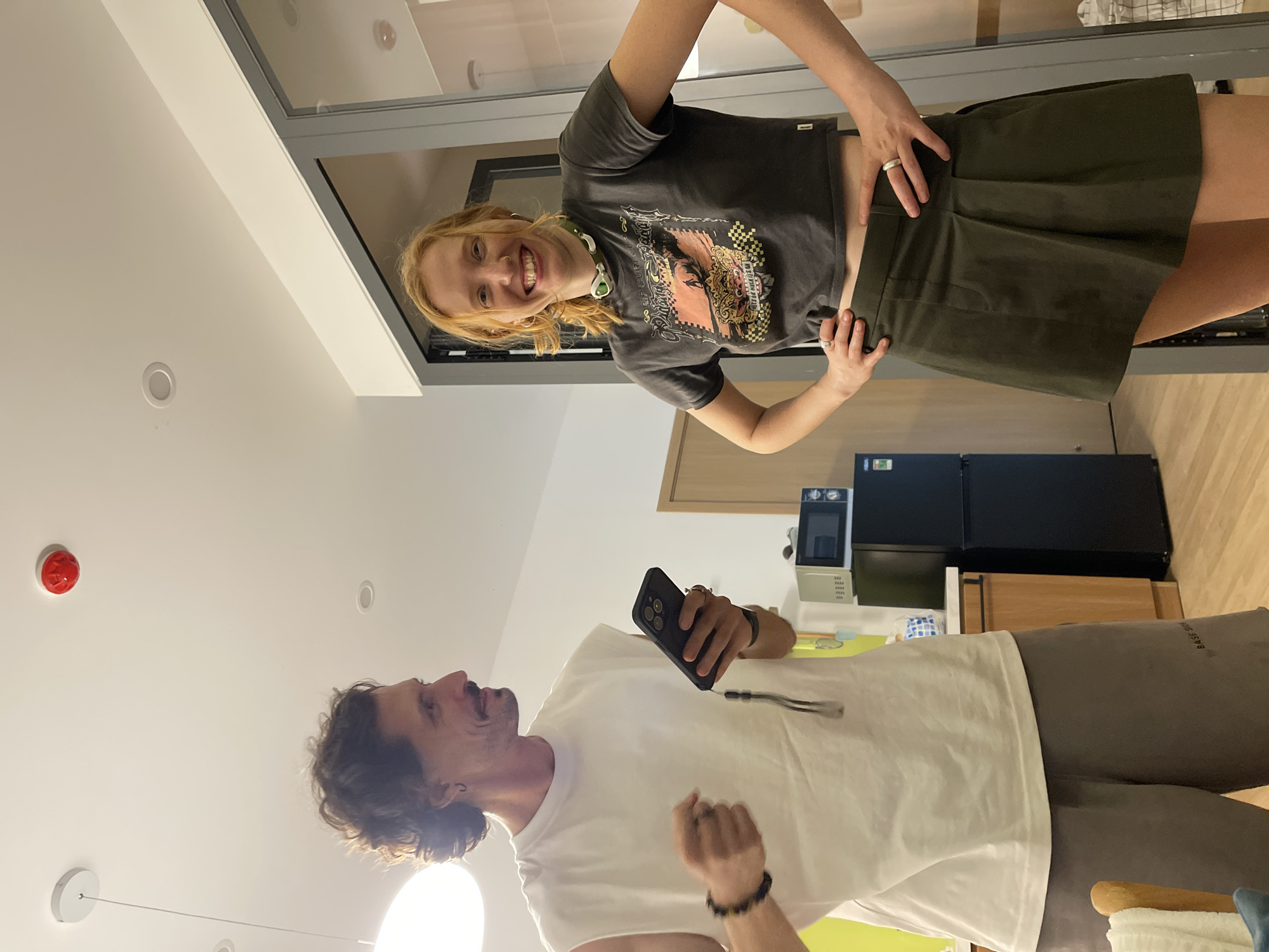 In a domestic interior setting, potentially a kitchen or living area, a man and a woman stand facing the camera. The man, on the left, has curly dark hair and a mustache. He is wearing a white sleeveless t-shirt and grey shorts. He holds a black smartphone in his right hand and wears a beaded bracelet on his left wrist. The woman, on the right, has red hair pulled back and is smiling. She wears a dark t-shirt with a graphic print and a dark green skirt. She has her hands on her hips and wears two rings. The room appears to be well-lit, with overhead lighting and a visible smoke detector on the ceiling. A black refrigerator and microwave are visible in the background, along with wooden cabinetry and a wooden floor. The overall impression is that of a casual indoor gathering or interaction.
