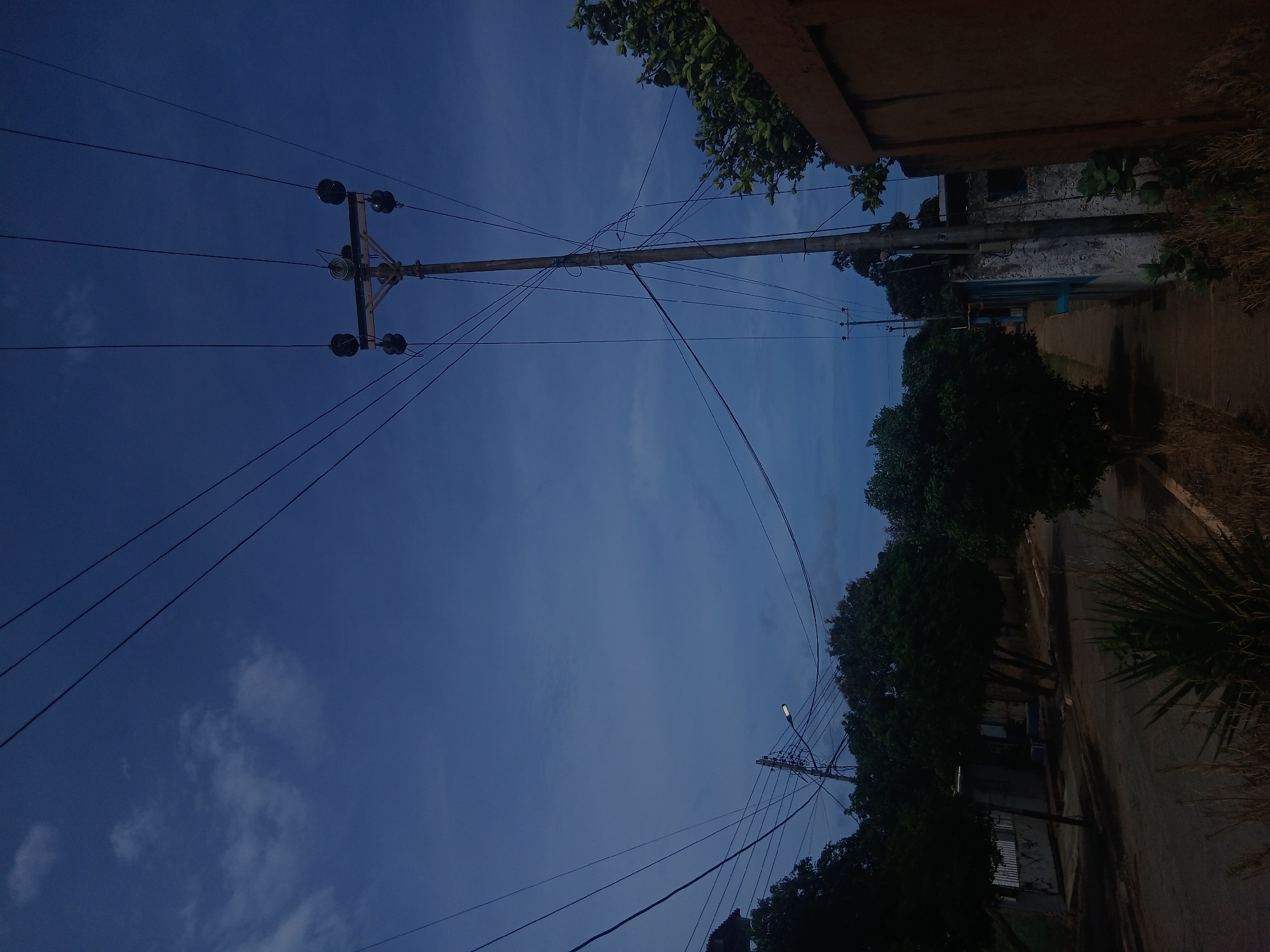 Looking upwards, the image shows a street scene in Juan Griego, Venezuela, with utility poles and power lines dominating the view against a blue sky with sparse clouds. A residential street with houses and trees lines the right side of the frame. The perspective is tilted. The time of day is late afternoon or early evening.