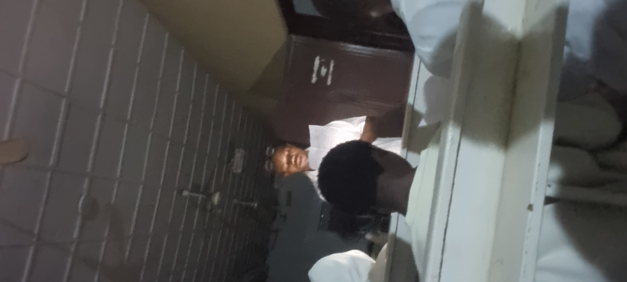 The image shows a dimly lit indoor scene, likely a classroom or meeting room in Lagos, Nigeria. In the center, a person wearing a white shirt and glasses stands holding a document, appearing to read or present. The ceiling is a suspended tile system with what seem to be ventilation fans. In the foreground, partially visible figures in white clothing are seated. A whiteboard with some markings is present on the right side of the frame. The overall atmosphere suggests an organized gathering or educational event.